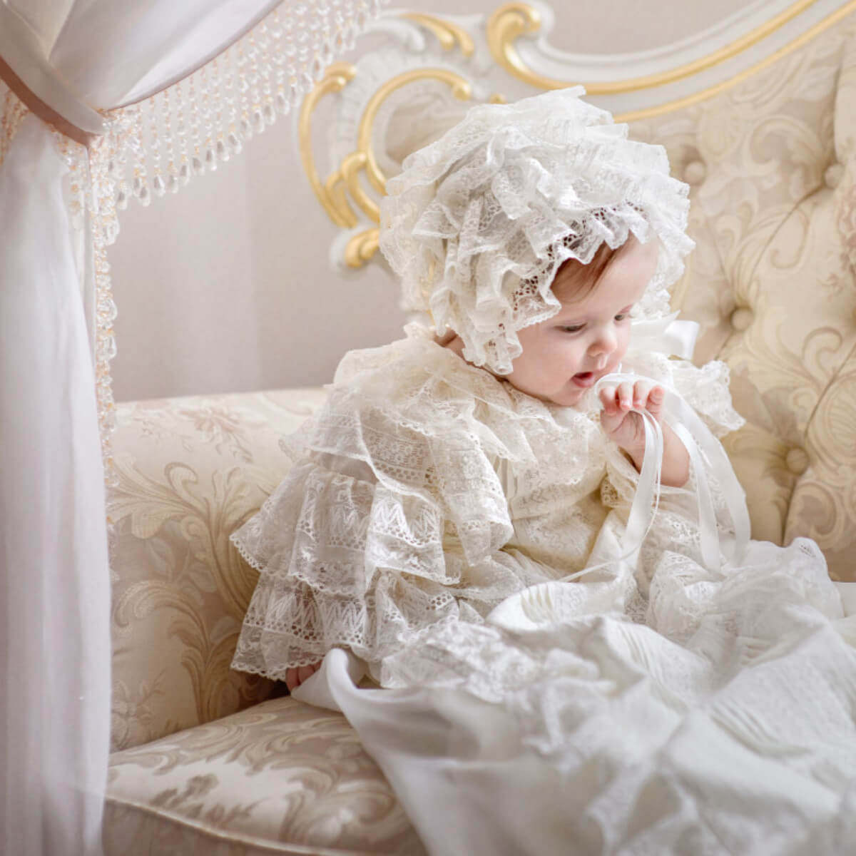 Baby in a white lace outfit sitting on an ornate chair