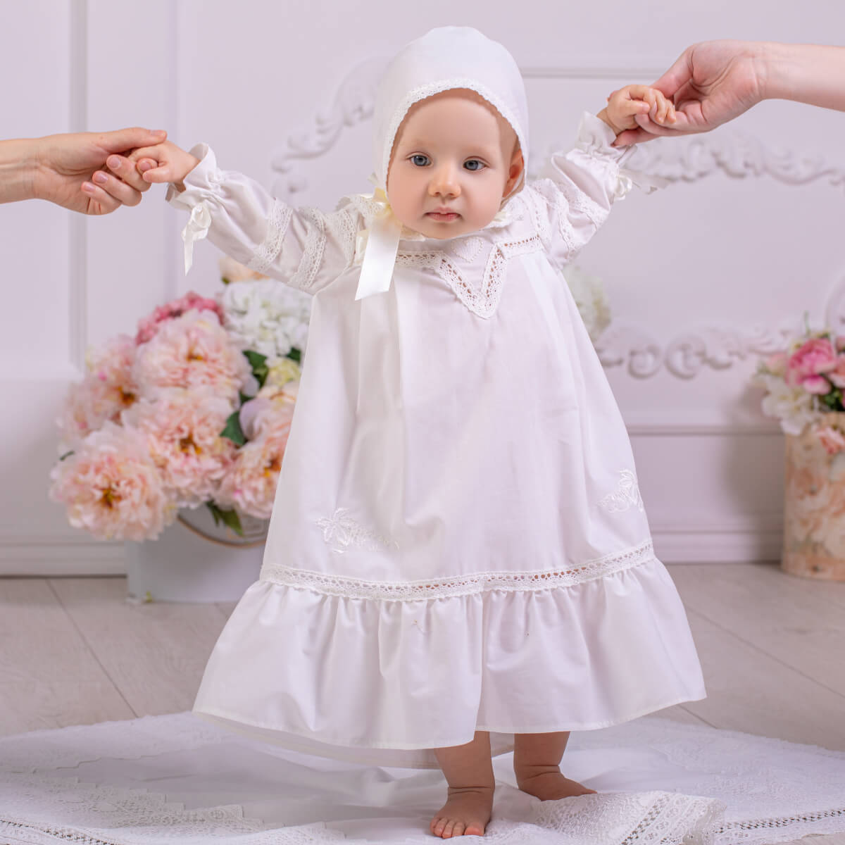 Baby in a Girls Baptism Gown standing on a white rug with floral decorations in the background