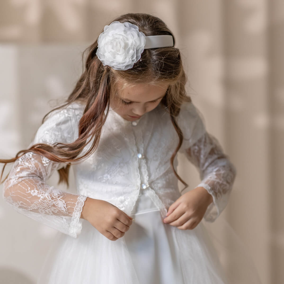 Young girl in a white lace Catholic Communion Dress with a floral headband against a neutral background
