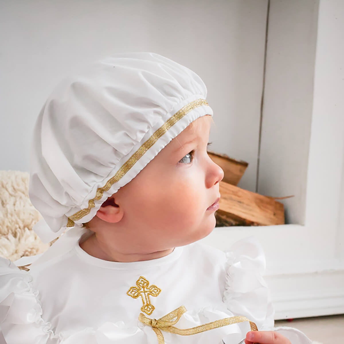 Baby wearing a Boy Christening Hat with gold trim and a white outfit with a gold cross design.