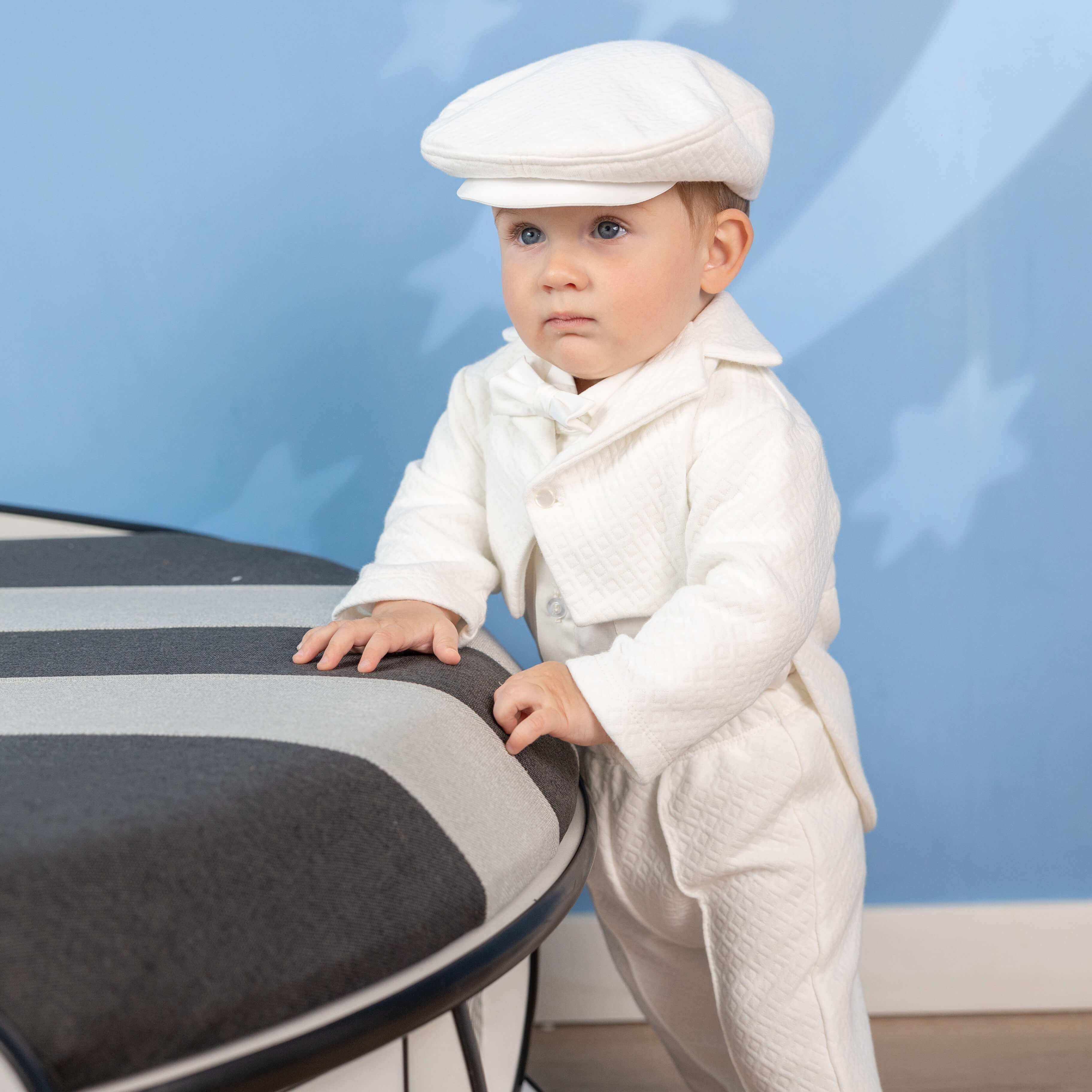 Baby in a Baby Boy Baptism Suit – with a cap standing next to a chair against a blue background