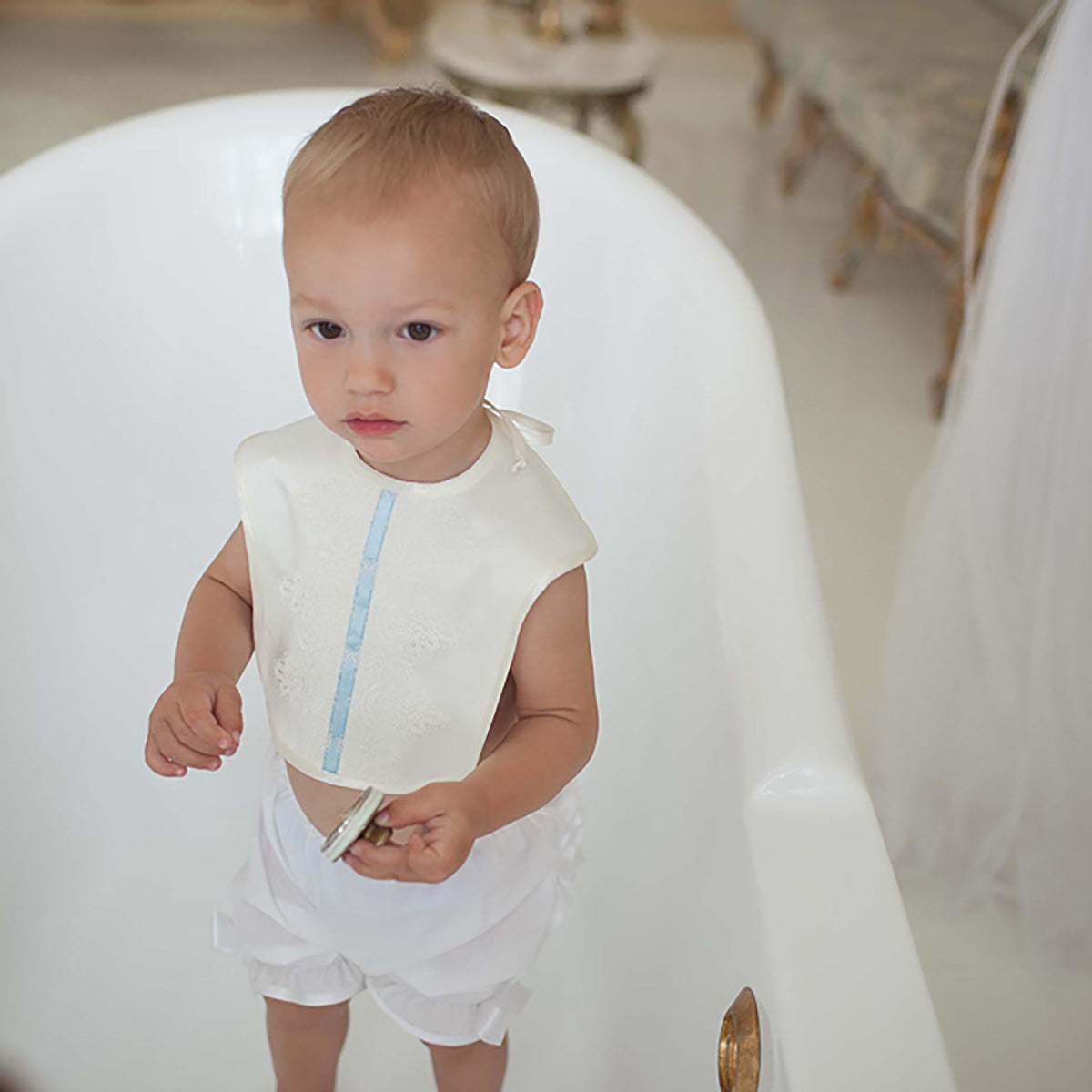 Child wearing a white outfit with a Baptism Bib in a softly lit room.