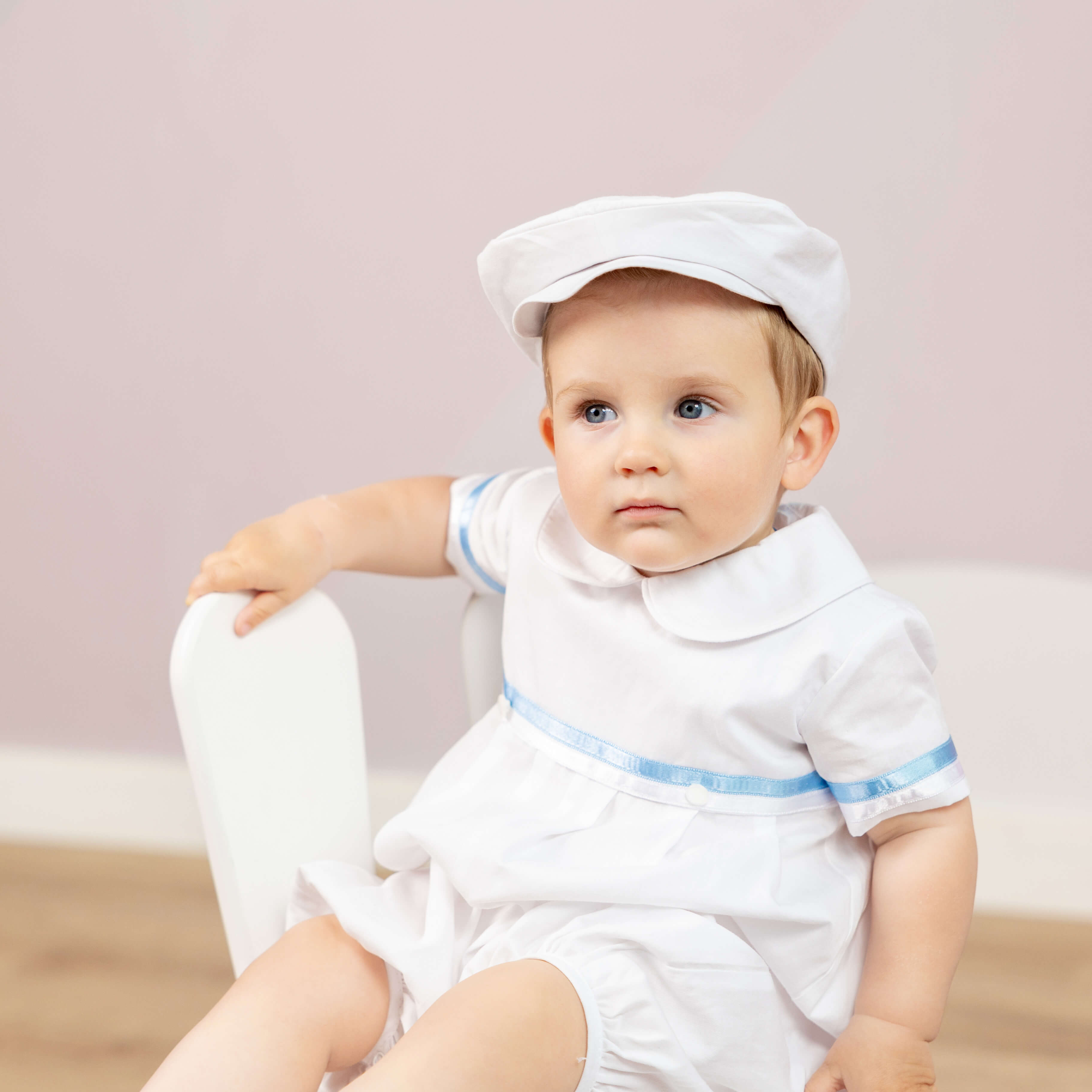 Baby wearing a white outfit with a blue trim and a Boy Christening Hat sitting on a chair against a light pink background.