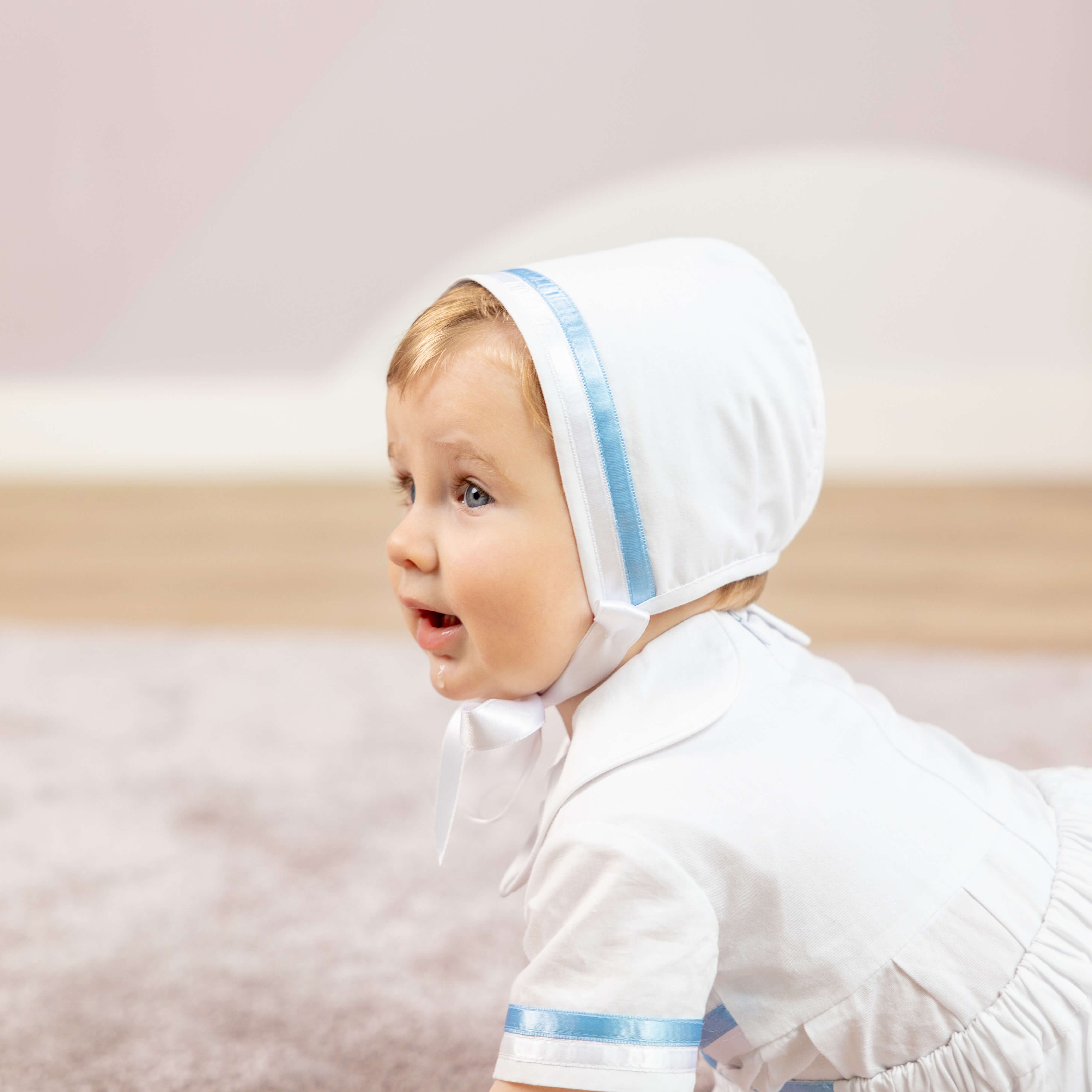 Baby wearing the Boy Christening Bonnet with blue trim on a light background