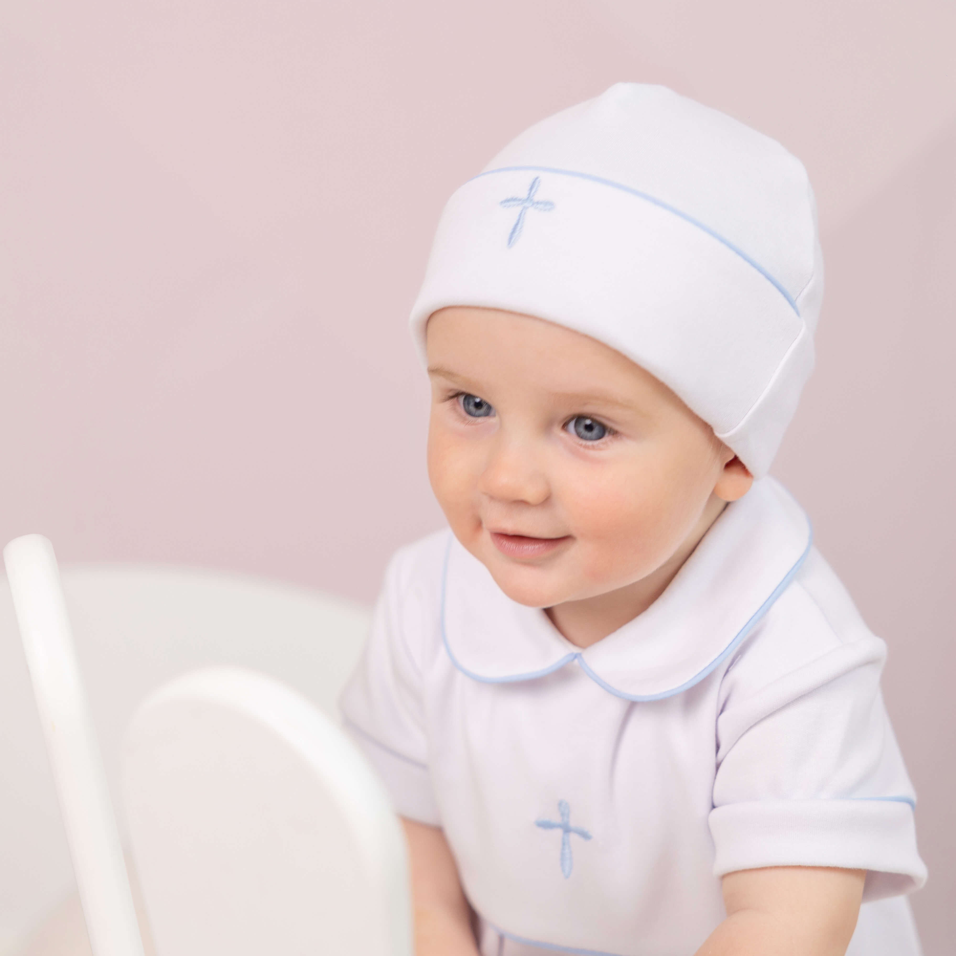 Baby wearing a Baptism Outfit for Boys with blue accents and a matching hat on a light pink background