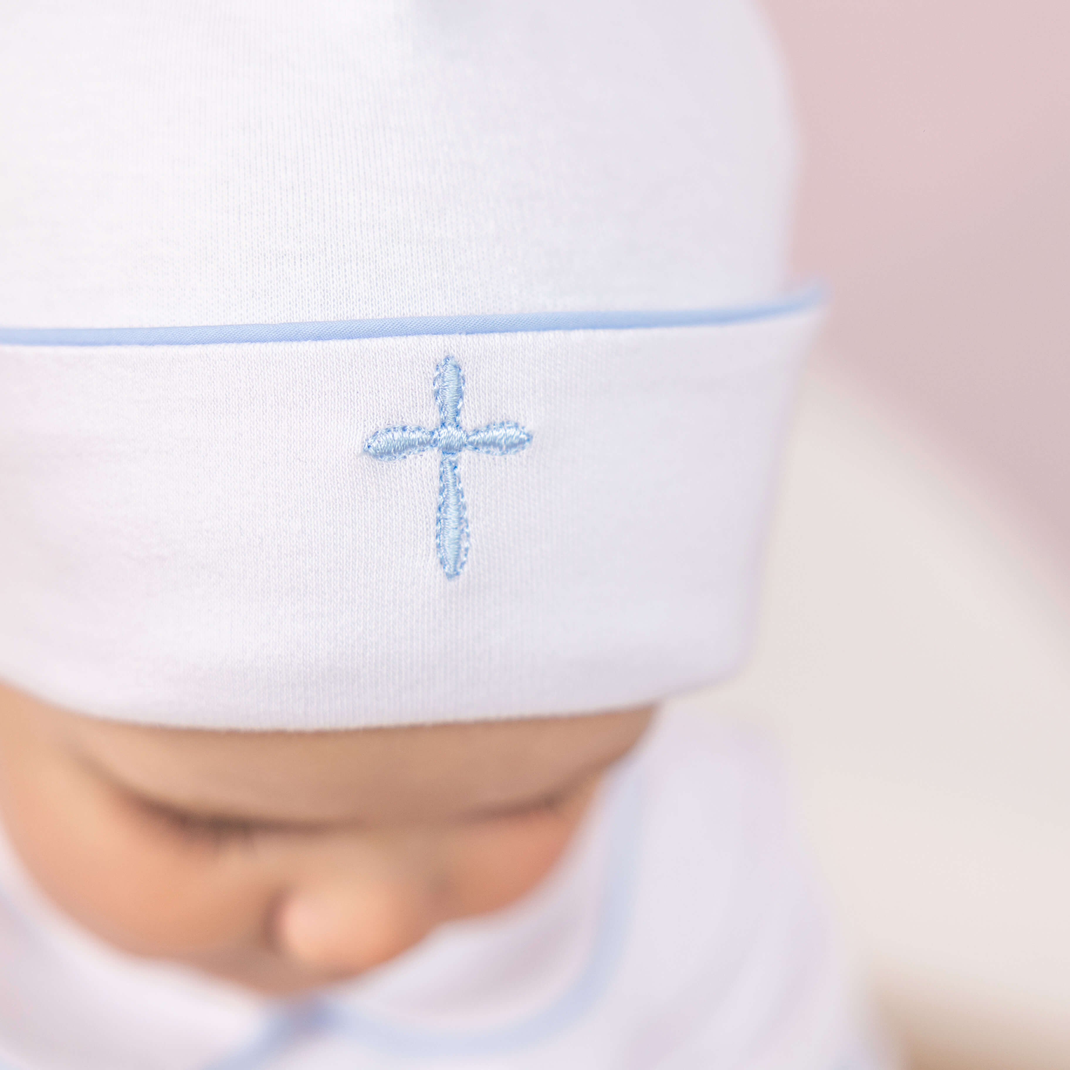 White baby hat with a blue cross of Baptism Outfit for Boys embroidery on a blurred background