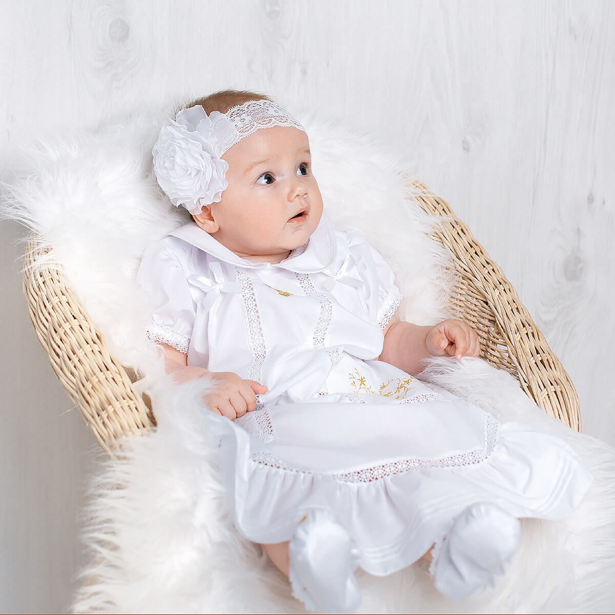 Baby in a White Christening Dress with lace details sitting on a fluffy white surface.