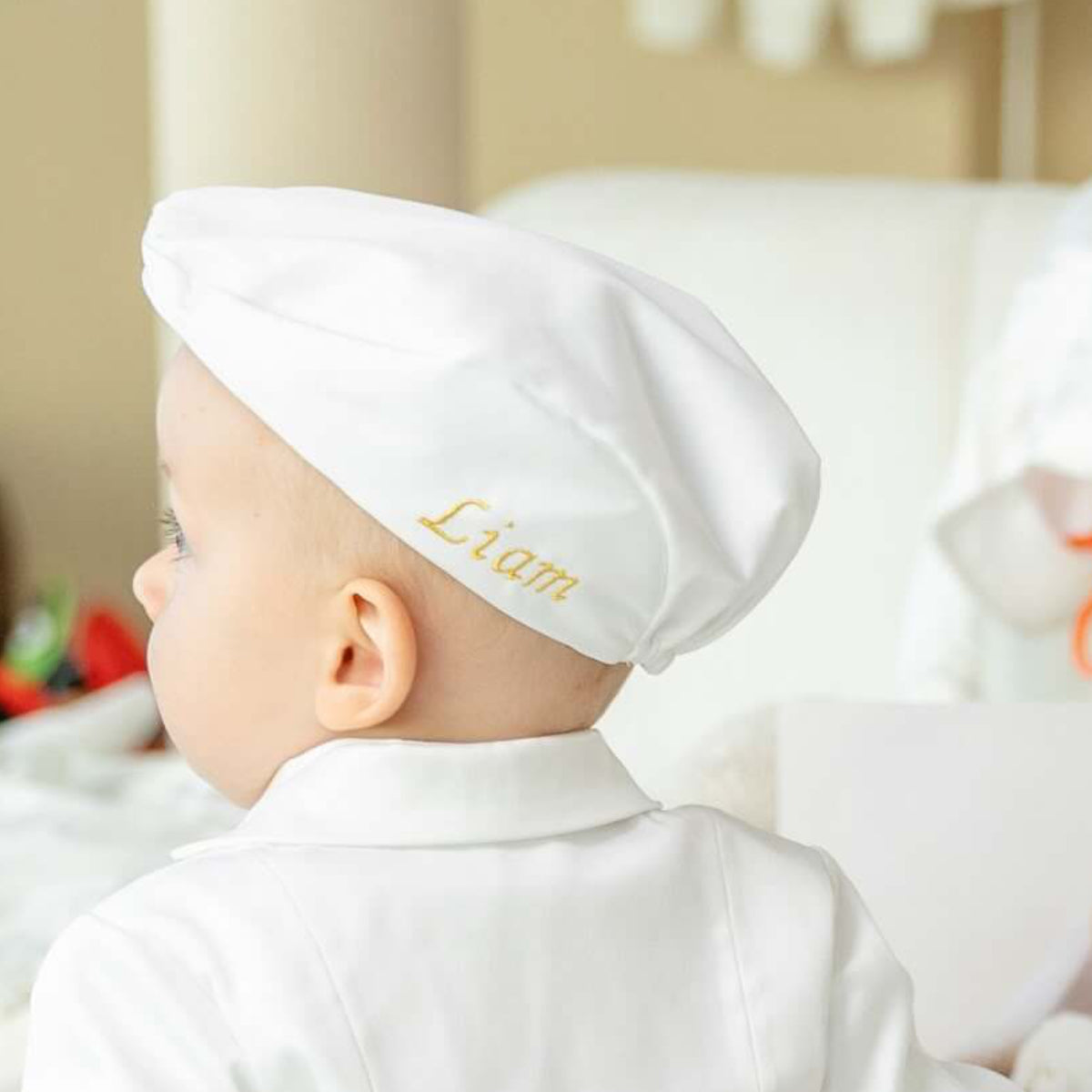 Baby wearing a Boy Christening Hat with 'Liam' embroidered on it, sitting on a couch.