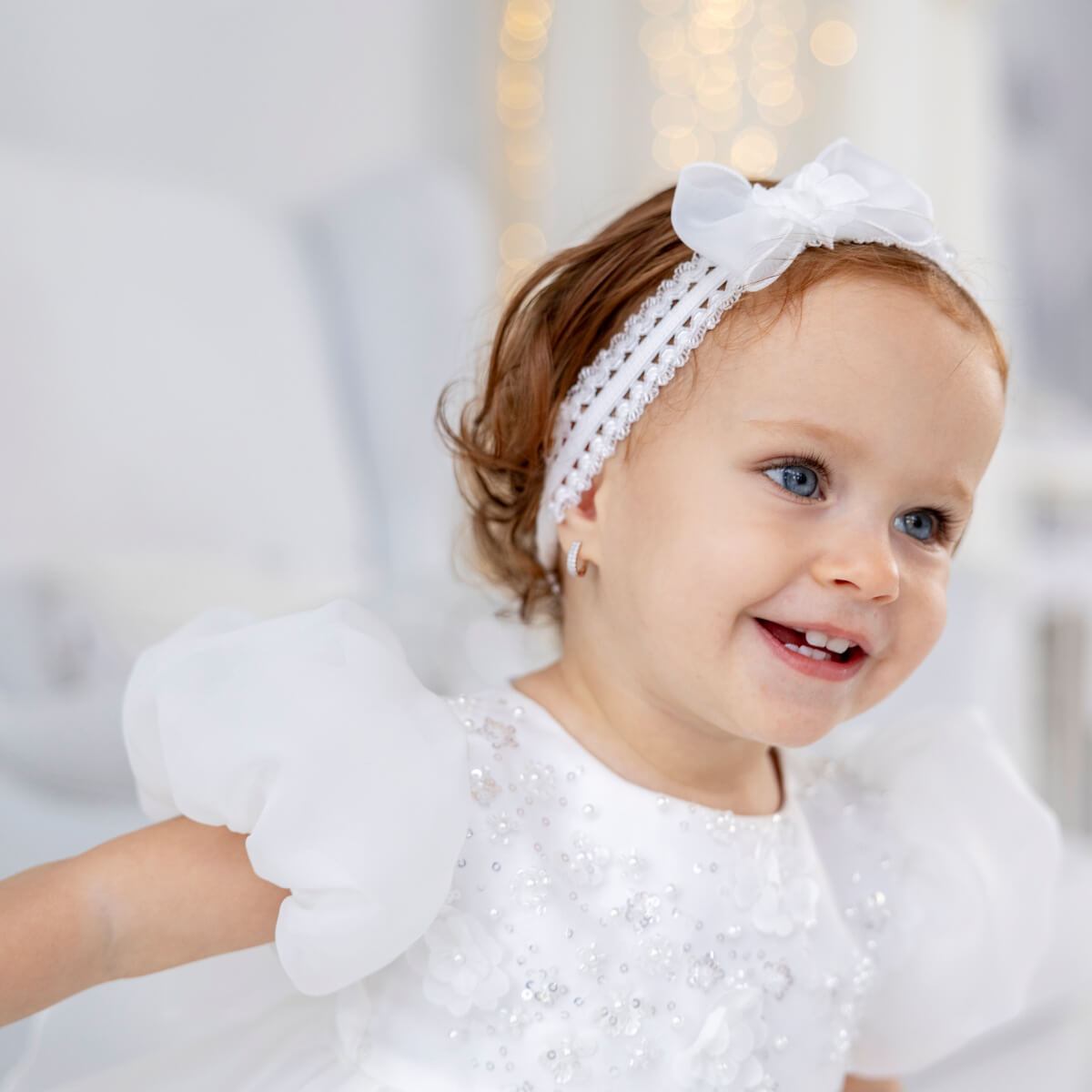 Girl in a white outfit with the baby headband, smiling against a blurred background
