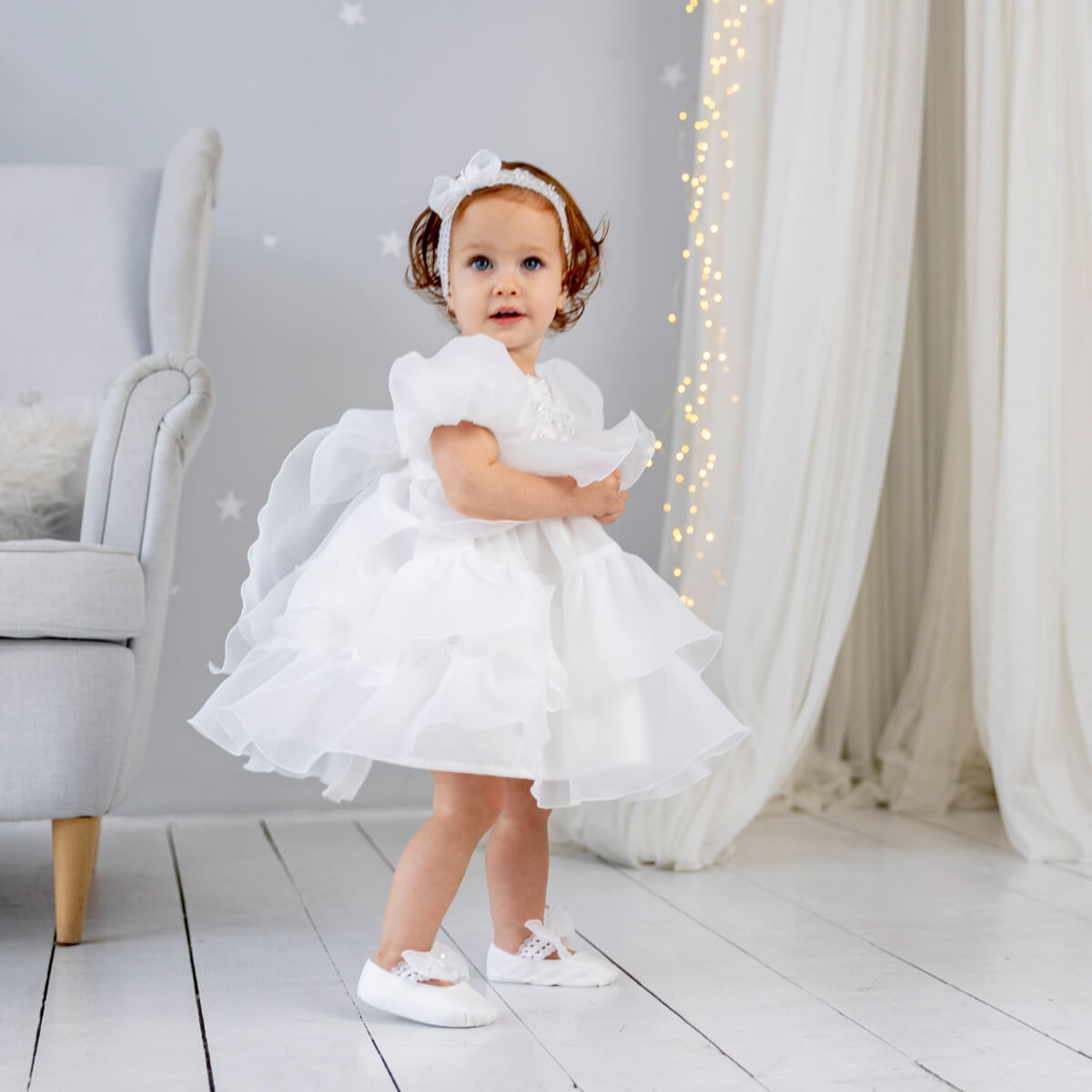 Young girl in a Baptism Dress with Detachable Train standing in a softly lit room with white furniture and curtains.