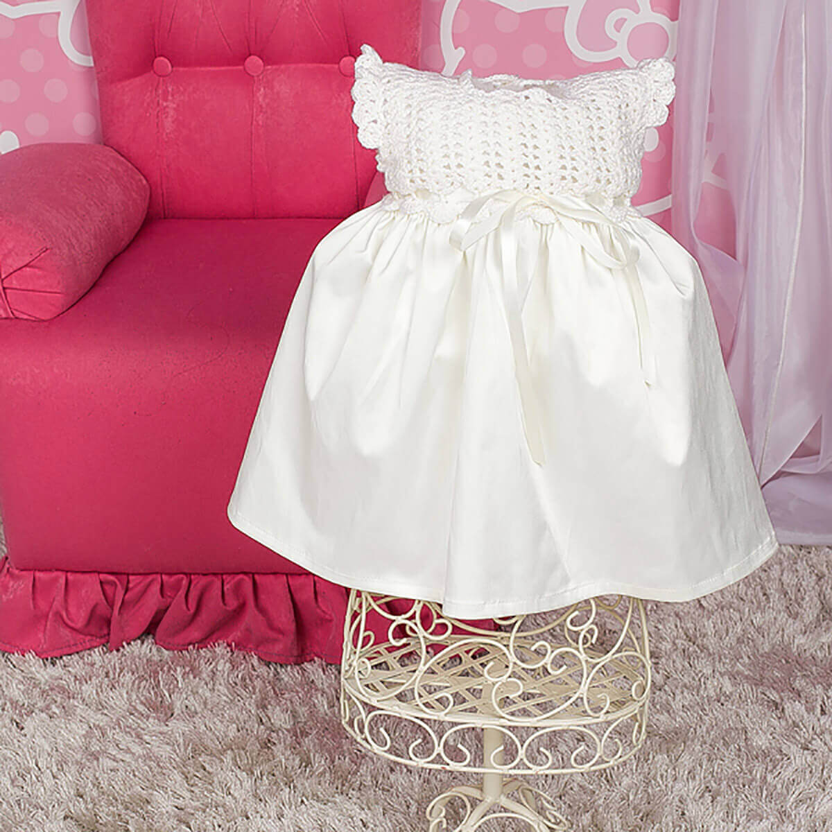 Crochet Christening Dress on a decorative stand in front of a pink chair with a polka dot pattern.