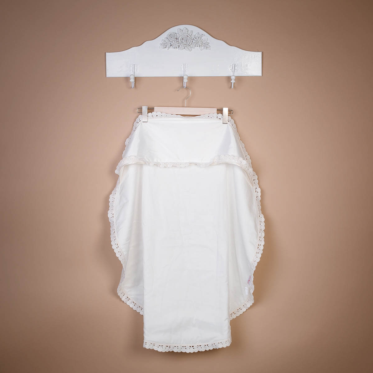 White lace baptism blanket hanging on a decorative wooden hanger against a beige wall.