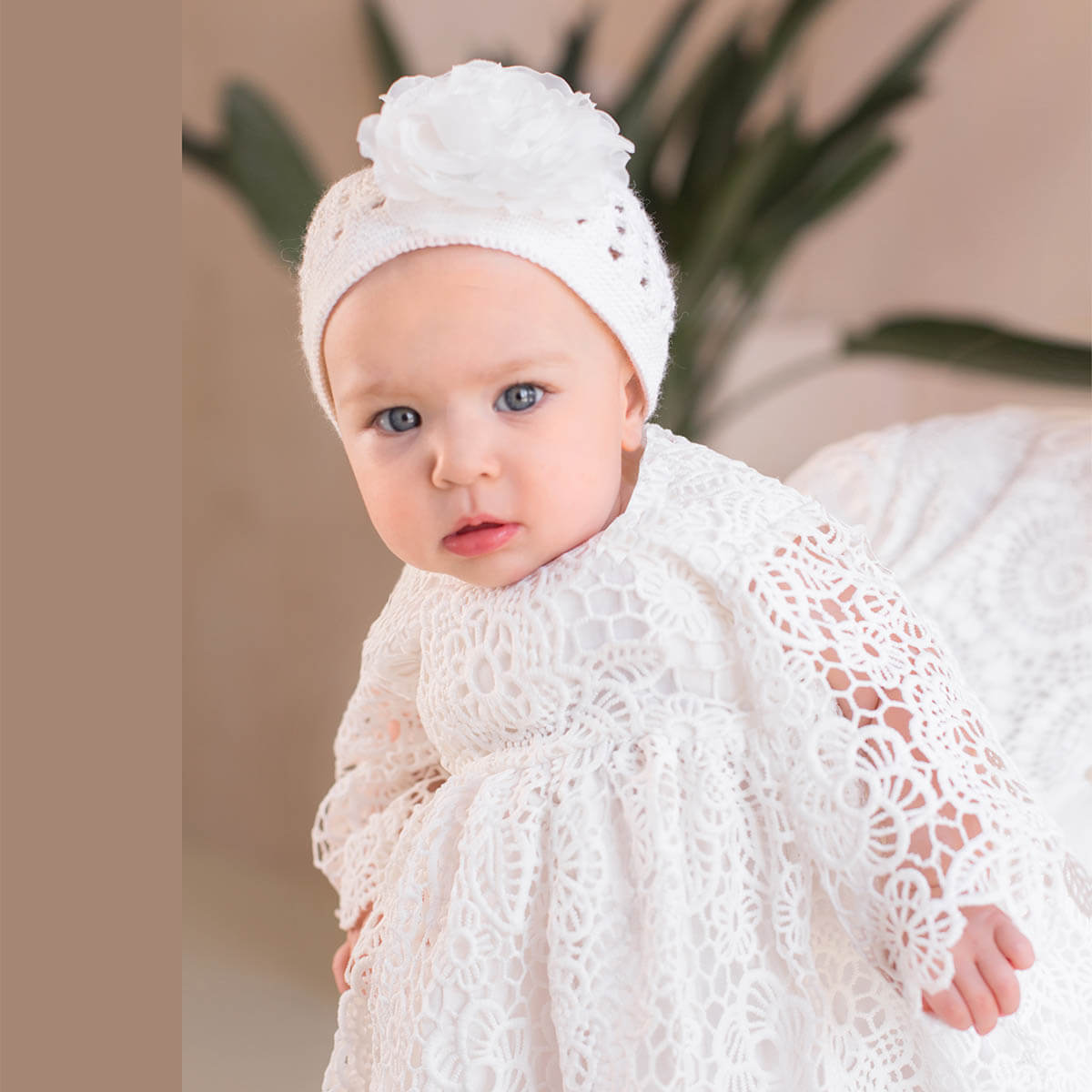 Baby in a white lace outfit with a crochet baptism bonnet against a blurred plant background
