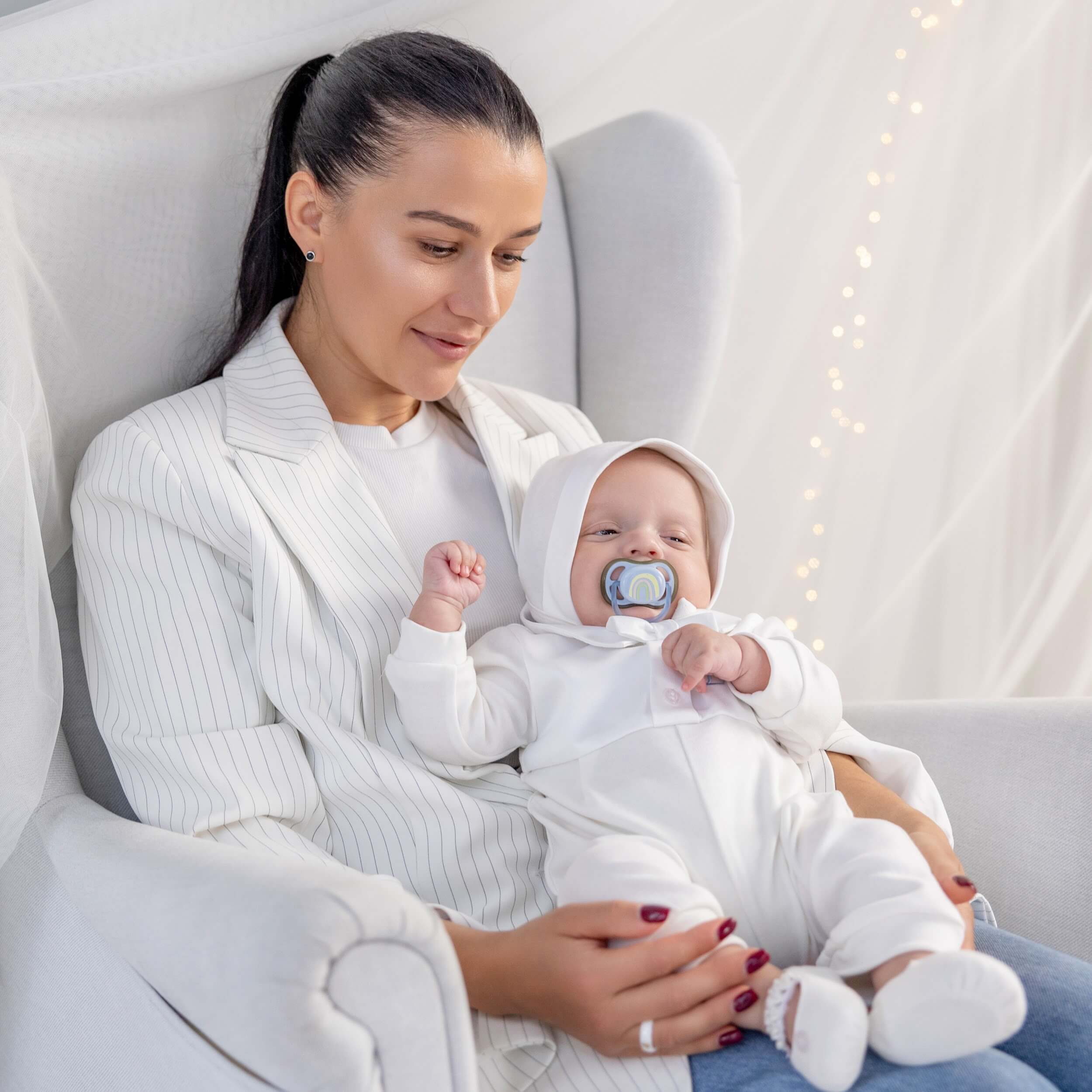 Woman holding a baby in a Coming Home Romper with a pacifier, sitting on a white couch.