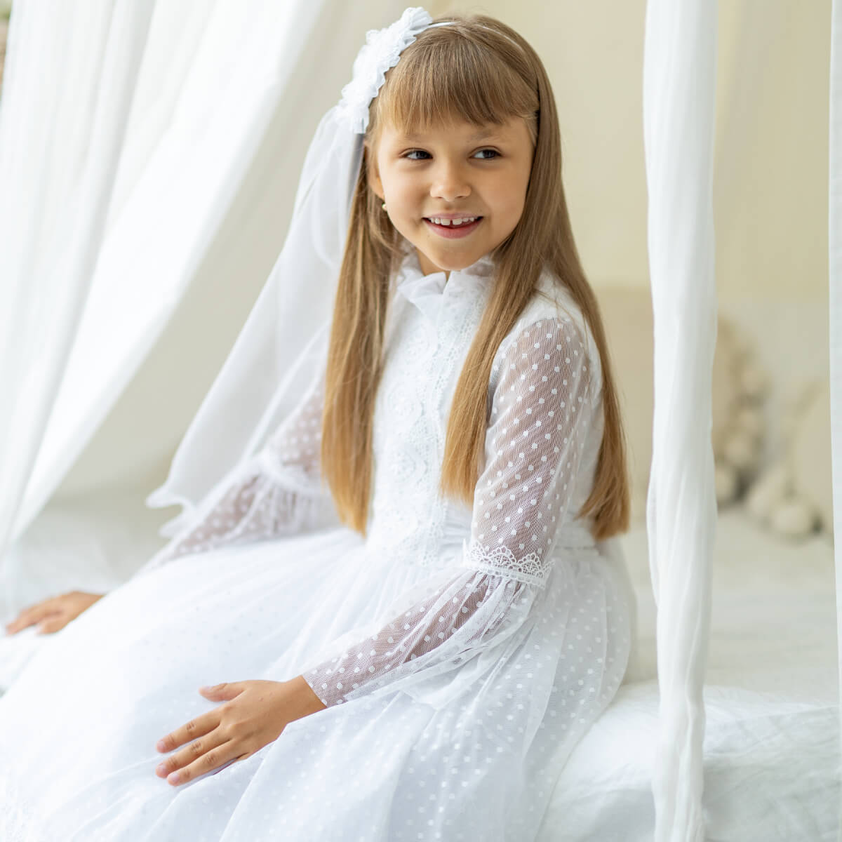 Young girl in a First Communion Dress with a veil sitting on a white couch.