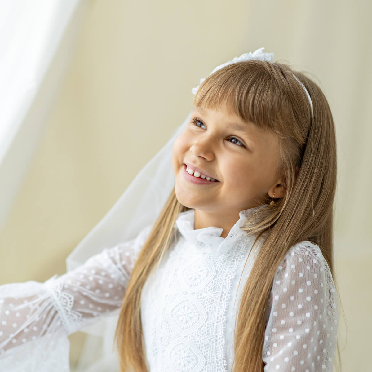 Young girl in a First Communion Dress with a veil, smiling indoors.