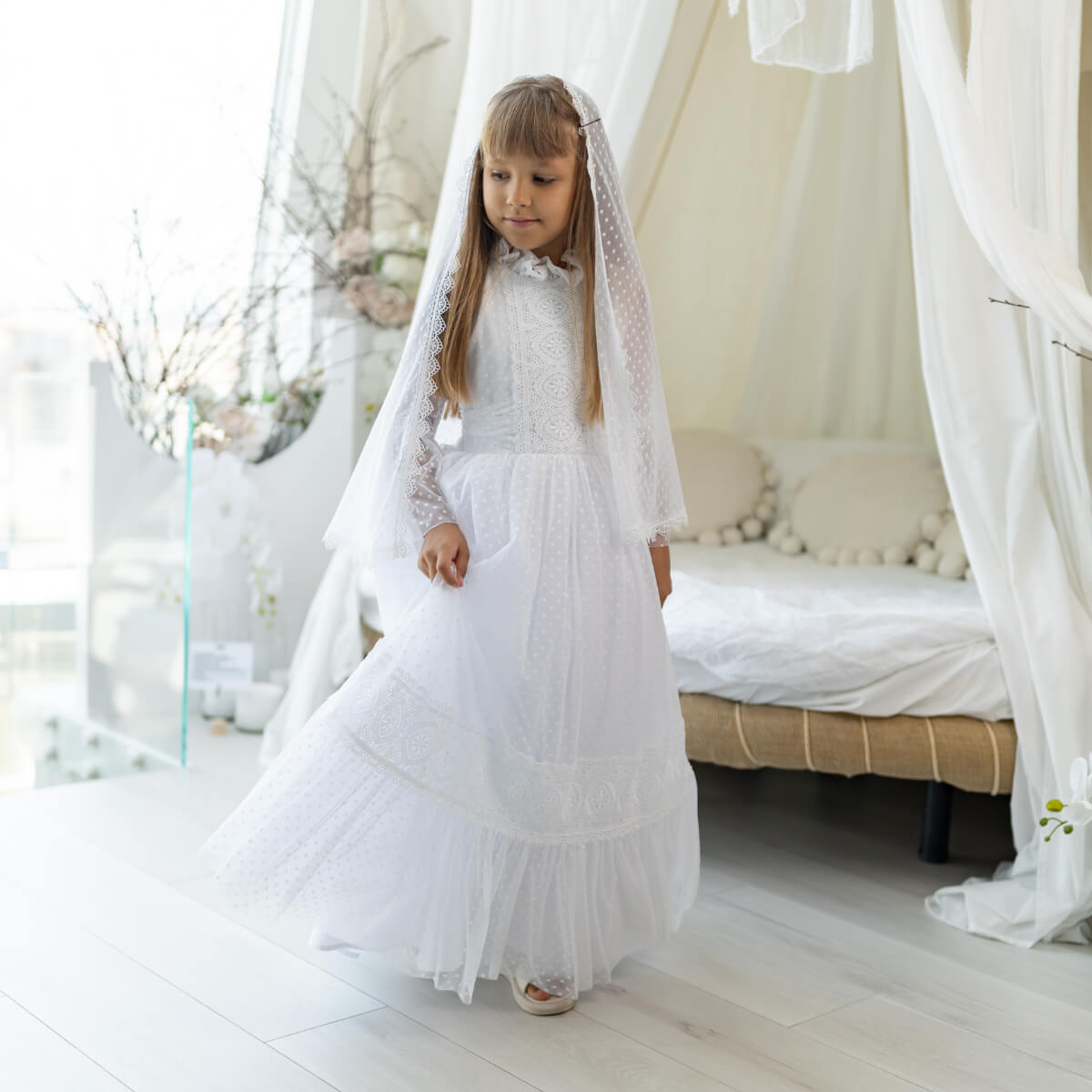 Young girl in a white dress standing in a softly lit room with white curtains and decor.
