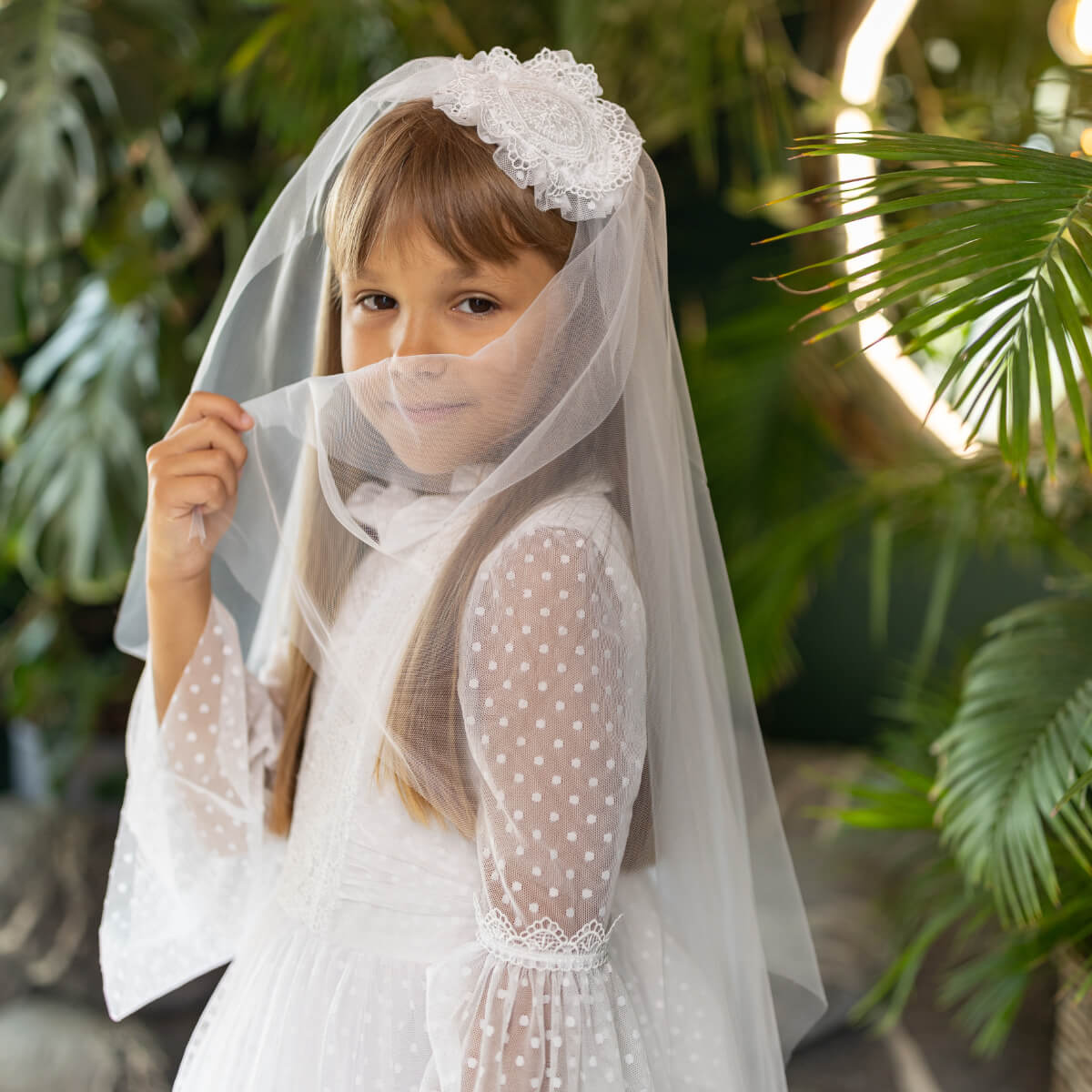 Young girl in a white dress with a First Communion Mantilla, standing among green plants.