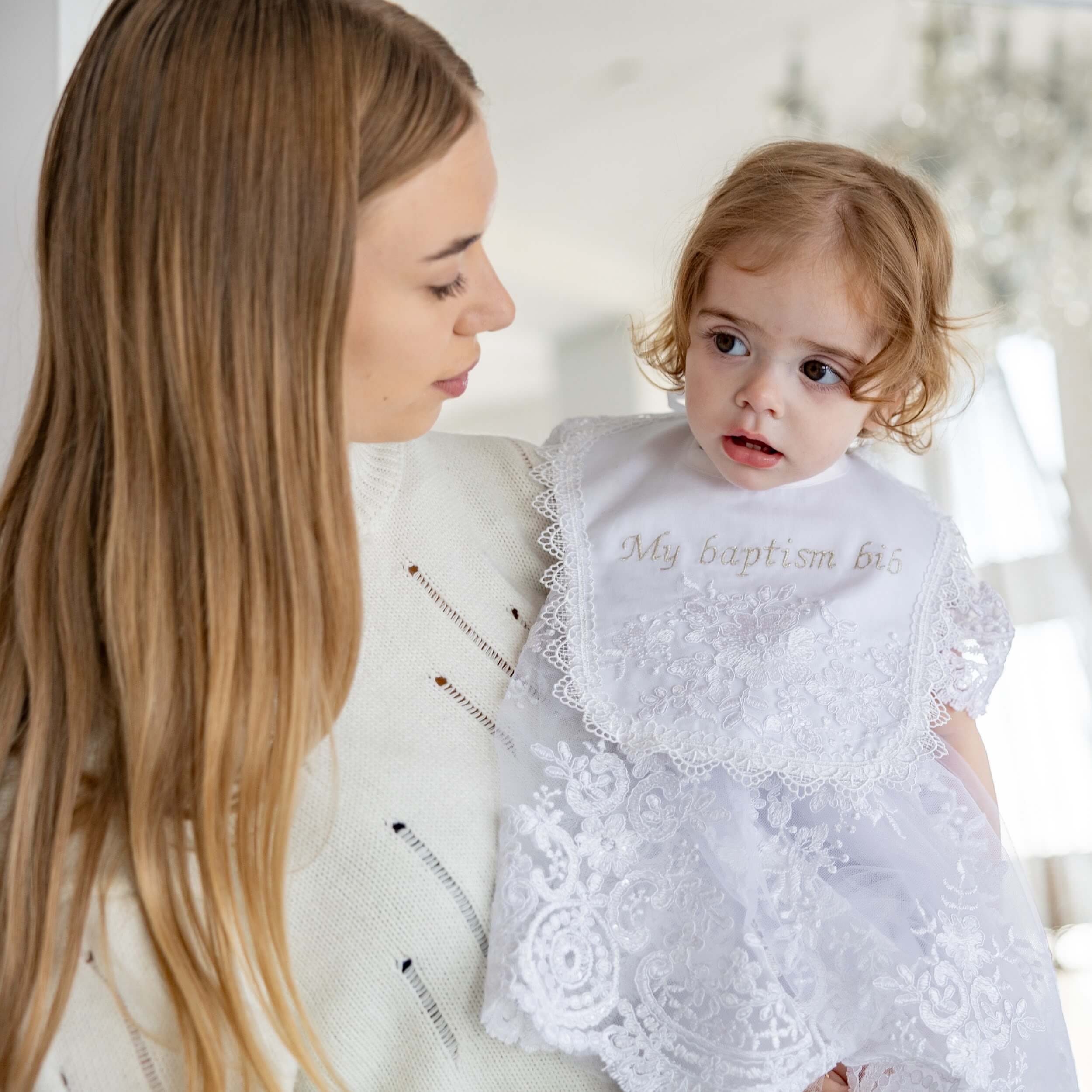 Woman holding a baby wearing a white lace outfit with bib 'My baptism bib' text.
