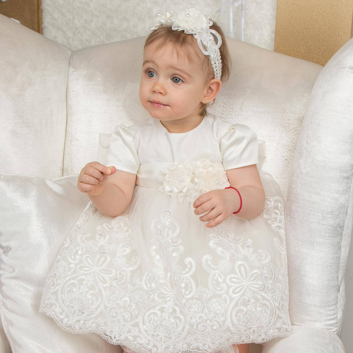 Baby in a Lace Christening Dress sitting on a white couch.