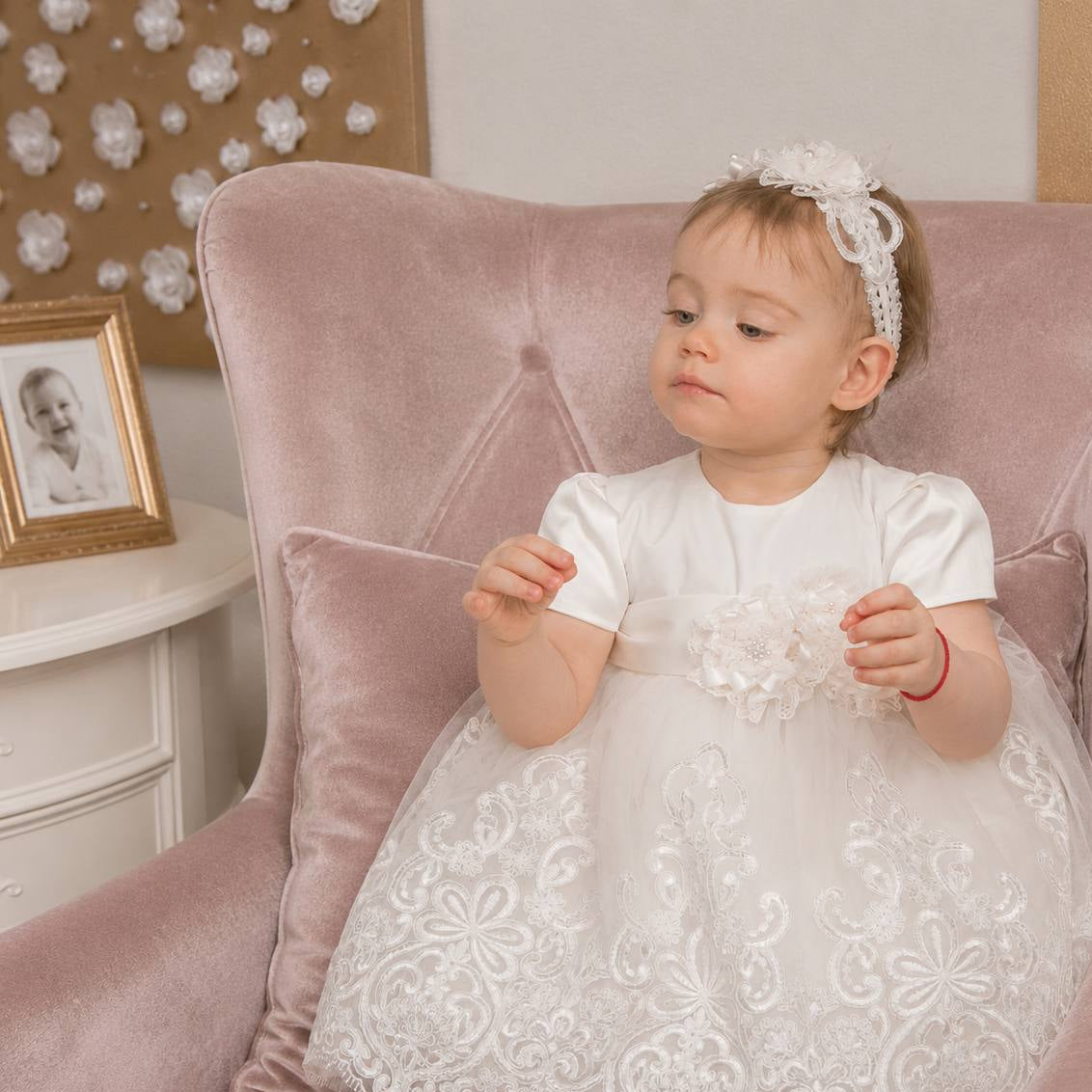 Baby girl in a white lace baptism dress sitting on a pink couch.