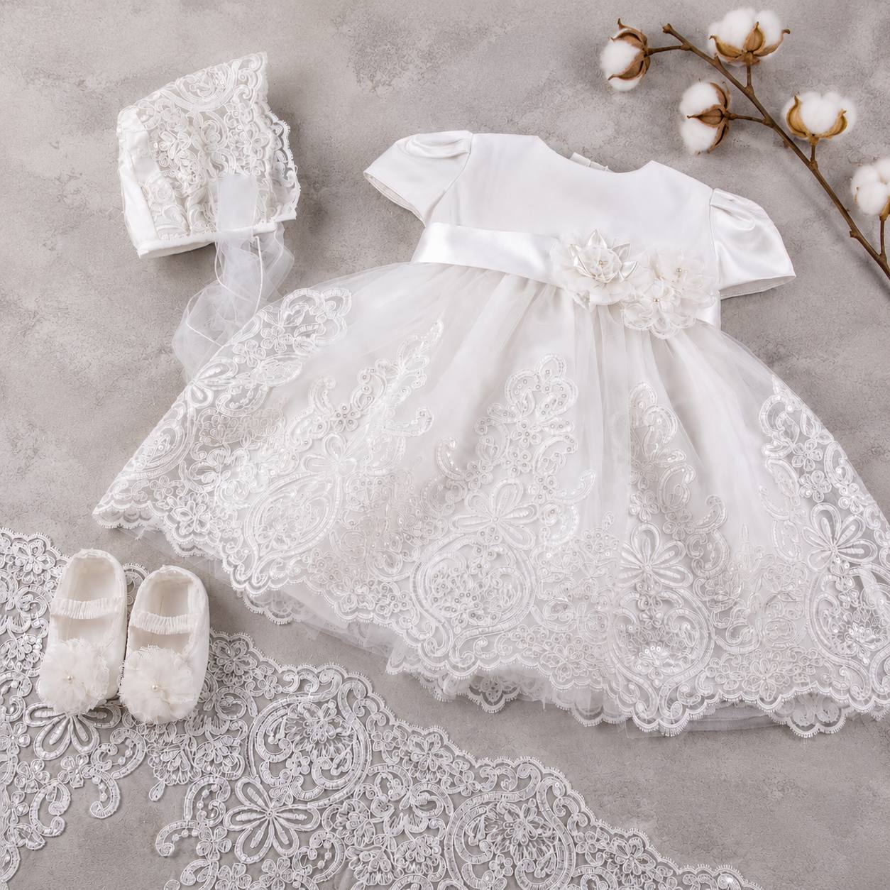 White lace baby dress with matching shoes and bonnet on a textured surface with cotton branches.