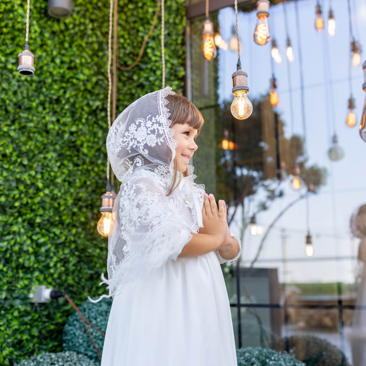 Young girl in a white dress with laHoly Communion Mantilla standing outdoors with decorative lights hanging above.