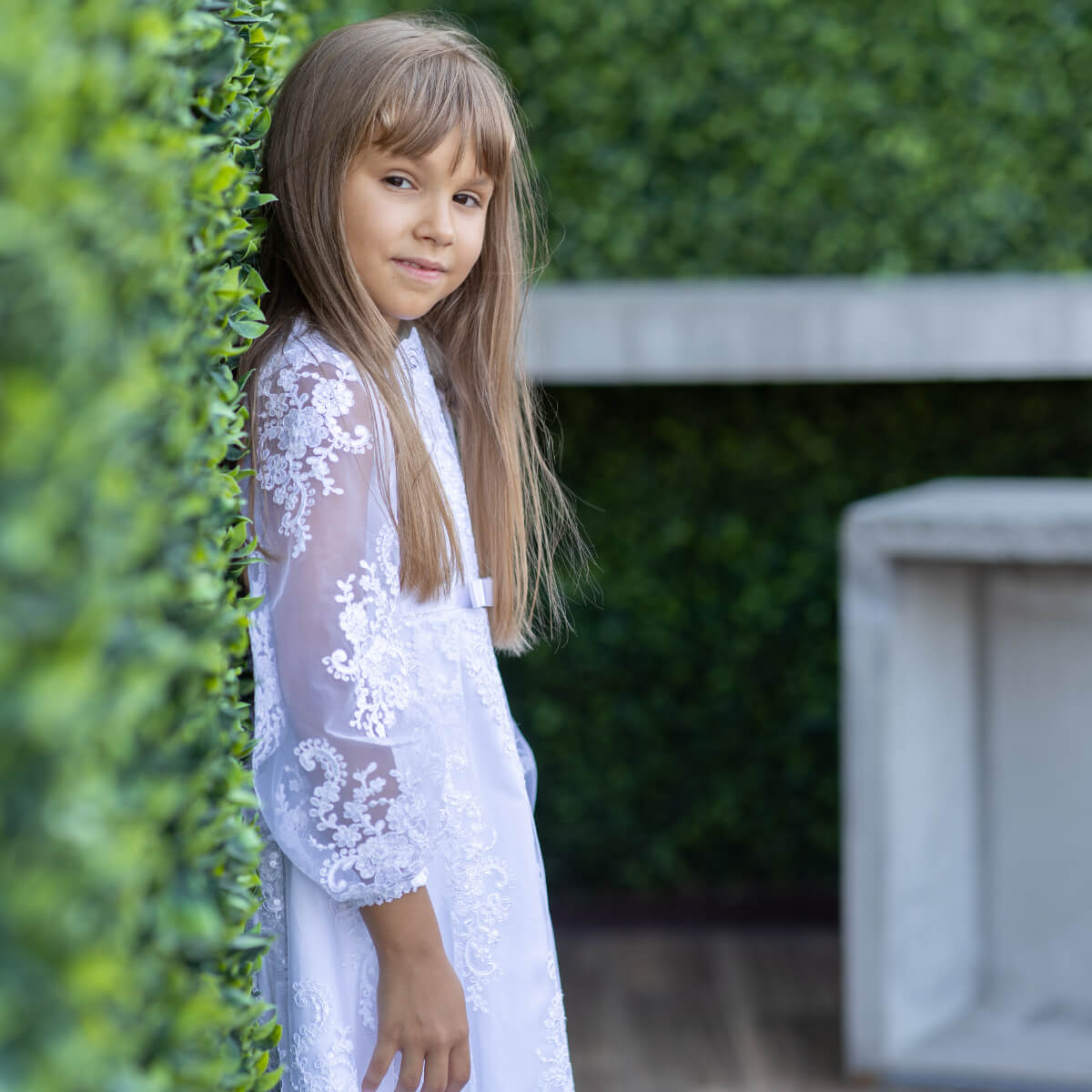 Young girl in a Simple Lace Communion Dress standing against a green hedge.