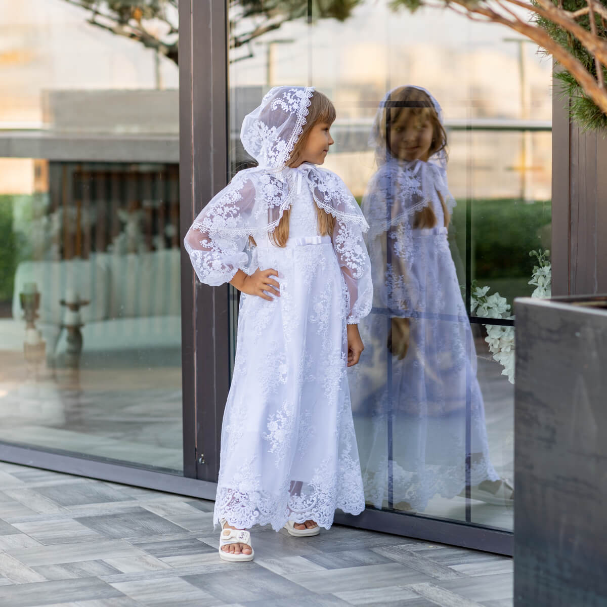Young girl in a white lace dress standing in front of a glass door.