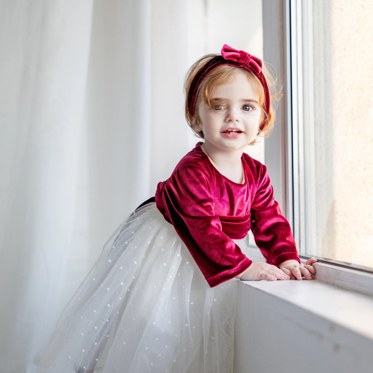 Young girl in a red top and white tulle skirt standing by a window.