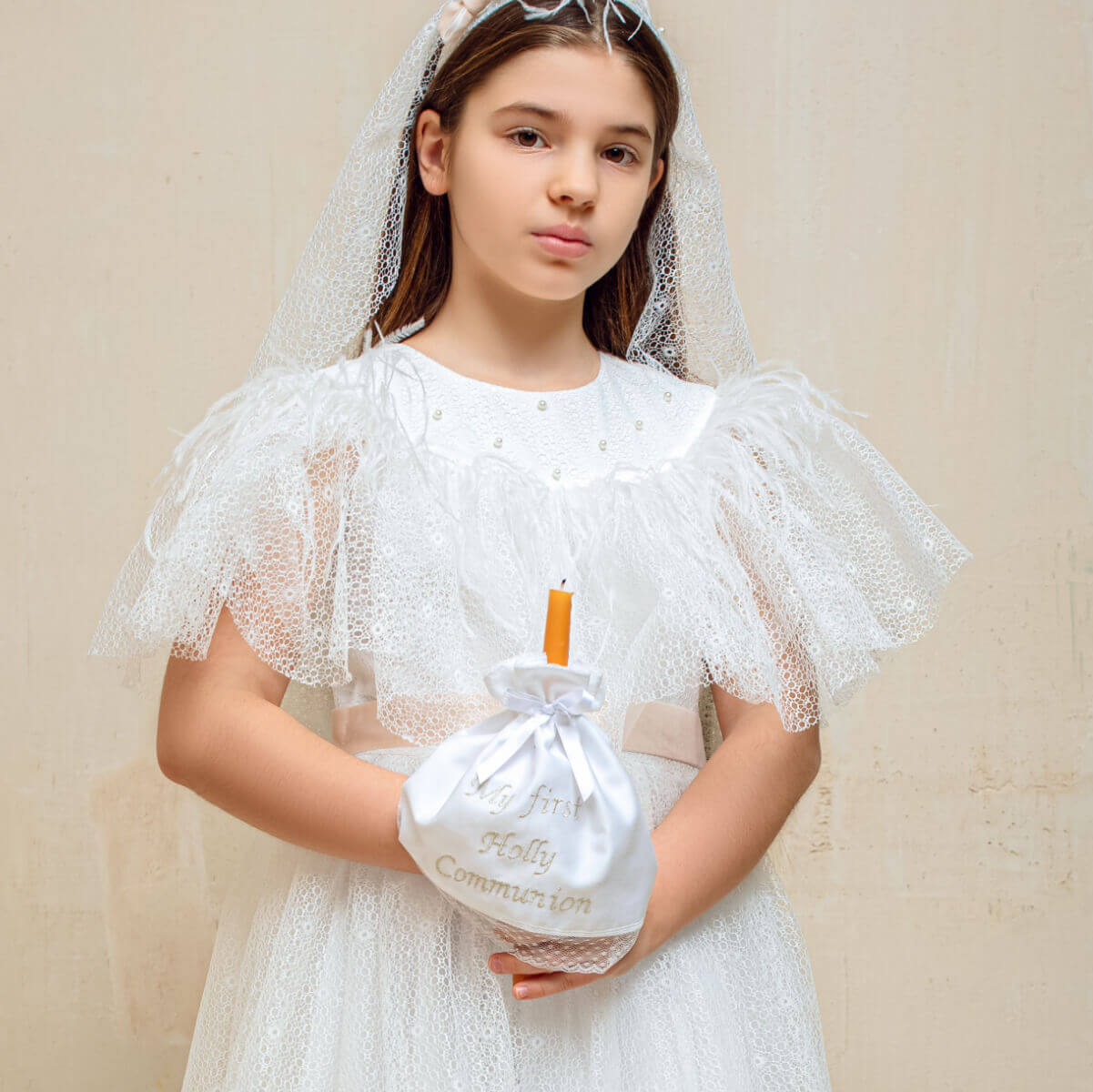 Young girl in a white communion dress holding a small bag with a candle, wearing a veil.