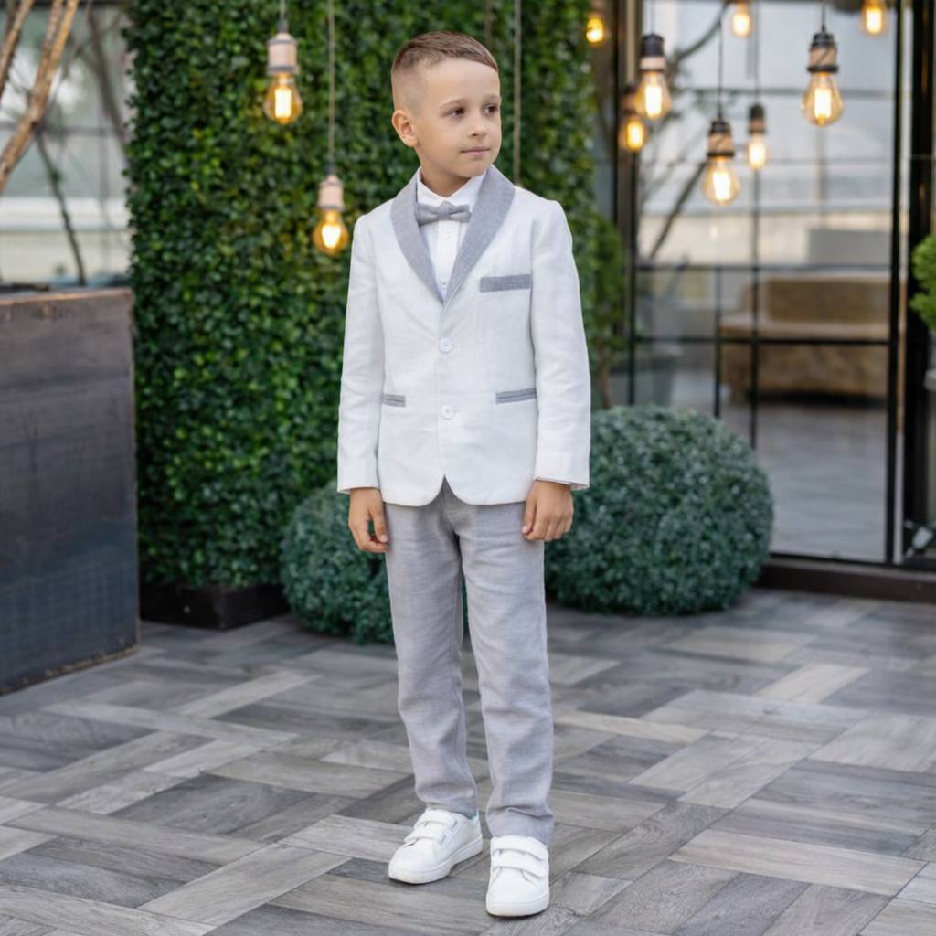 Young boy in a light gray suit standing outdoors with decorative lights and plants in the background.