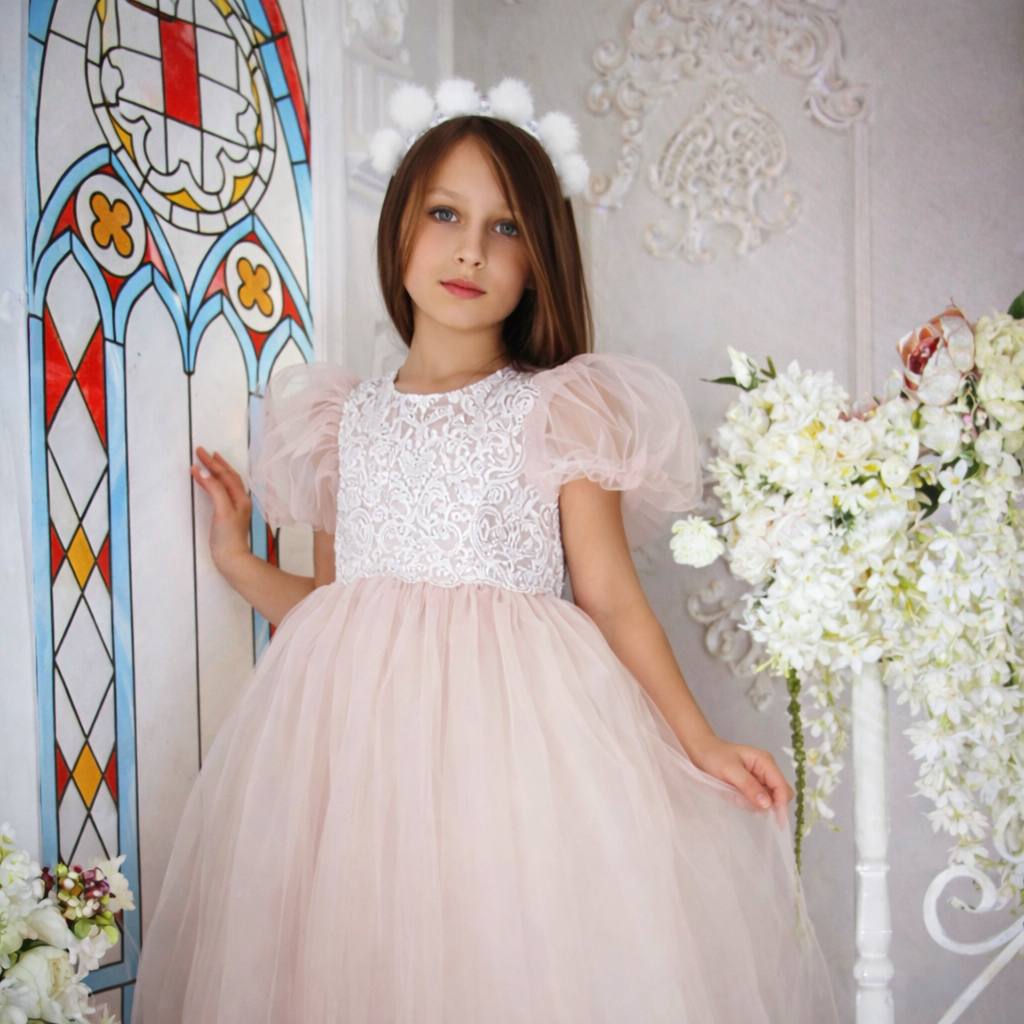 Young girl in a white lace top and pink tulle dress standing in front of a stained glass window and floral decorations.