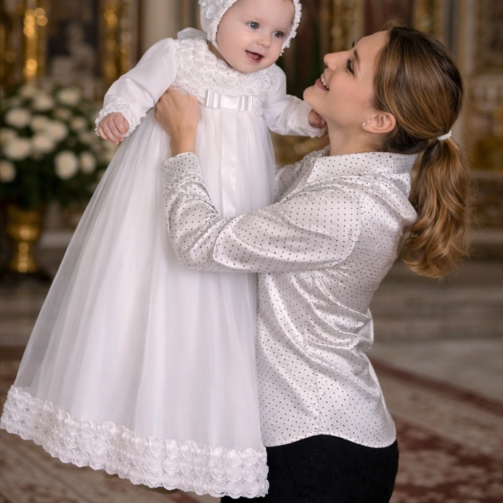Woman holding a baby dressed in a white outfit with lace details, in an indoor setting.