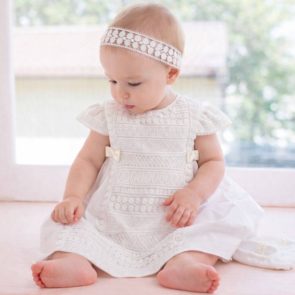 Baby in a white lace dress sitting on a light surface with a blurred natural background