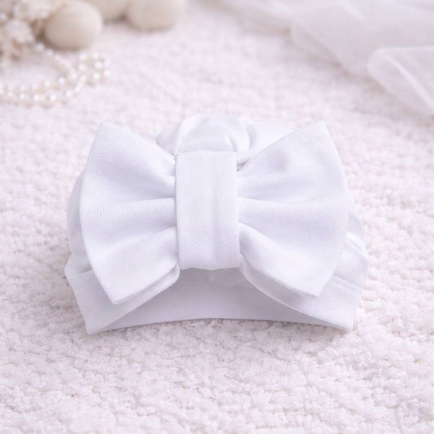 White bow headband on a textured white surface with a teddy bear and pearls in the background.