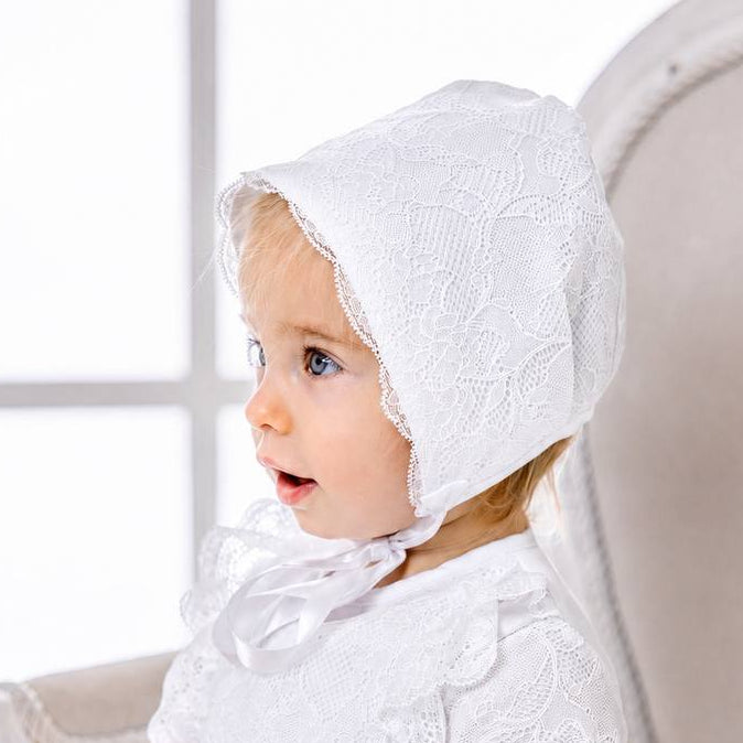 Baby in a white dress and bonnet sitting on a white chair.