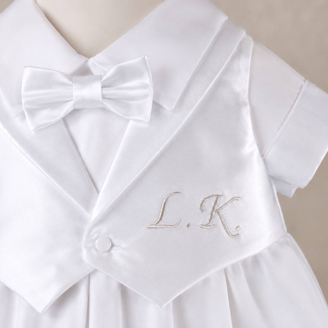 White baby outfit with a bow tie and 'L.K.' monogram on a beige background