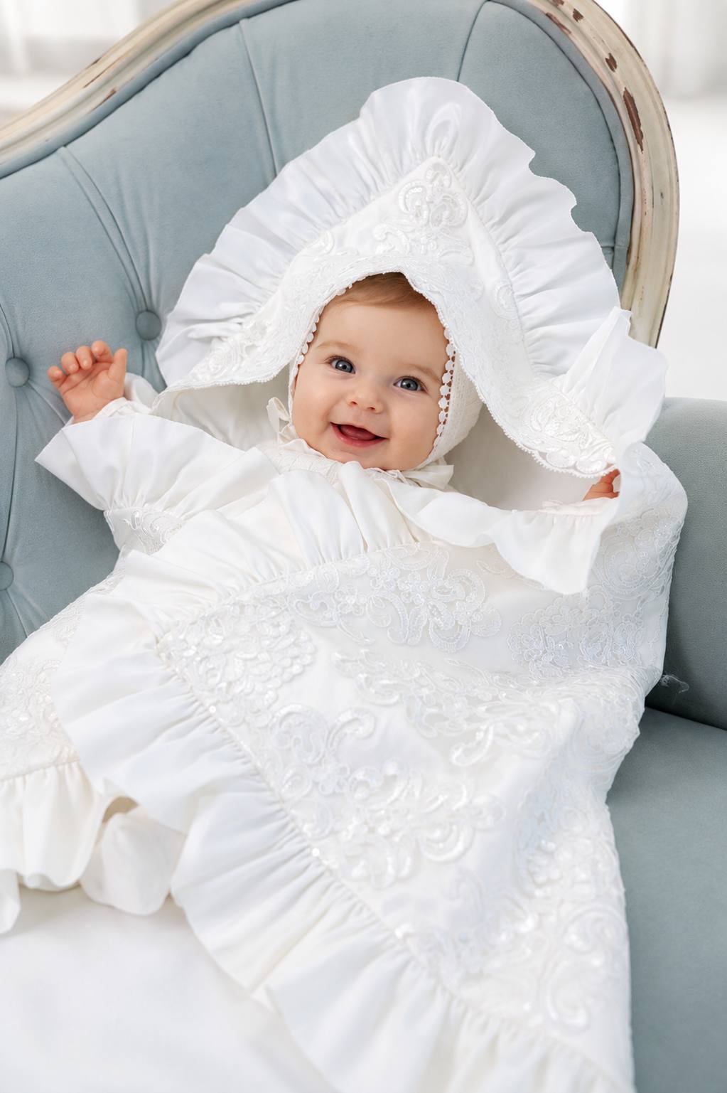 A baby wearing a white embroidered montera dress with a matching ruffled bonnet, lying on a christening blanket.