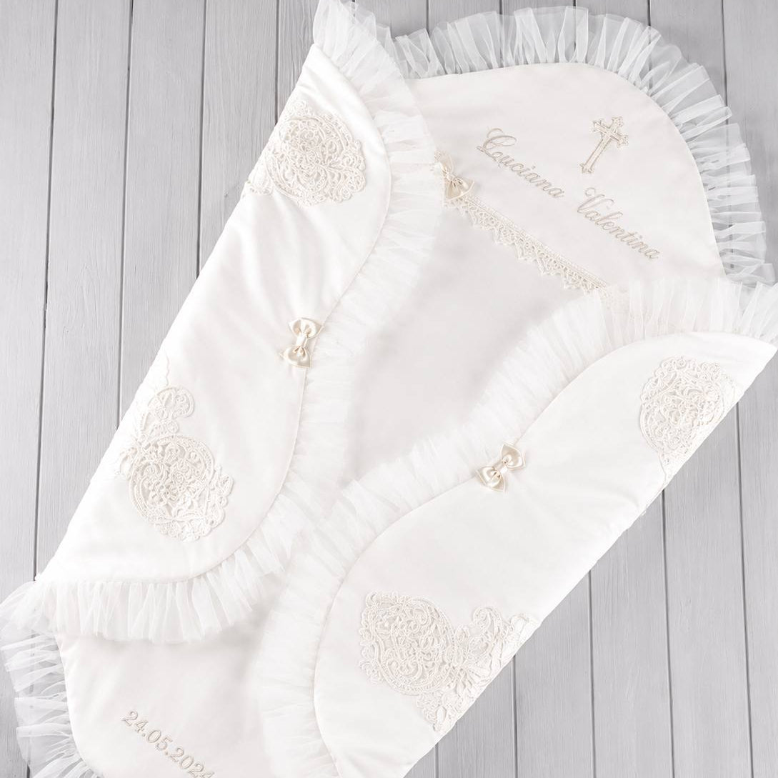 A white personalized baby swaddle blanket with delicate ruffles, embroidered floral motifs, and a monogrammed hood, displayed on a light gray wooden plank background.