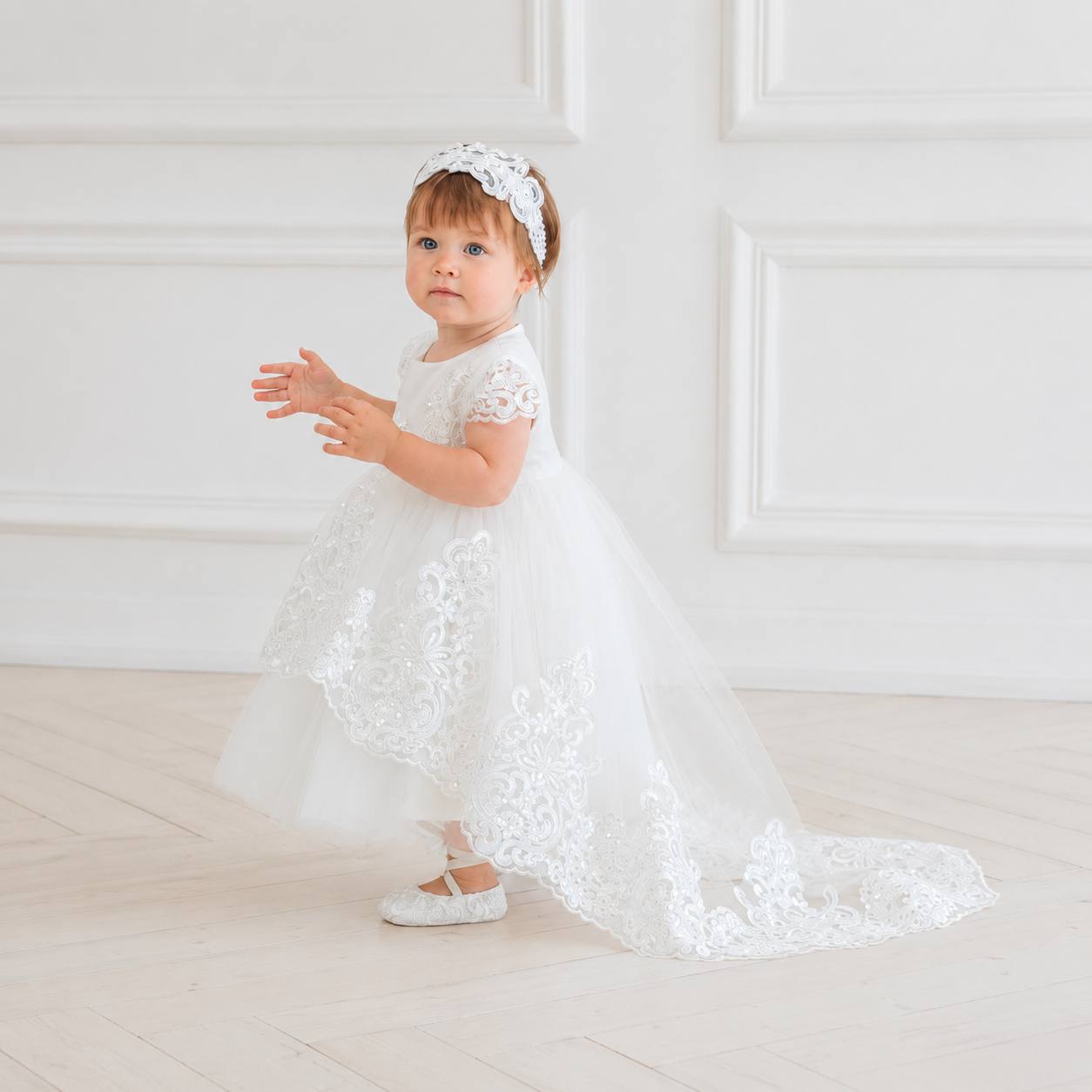A toddler girl wearing a white lace and tulle baptism dress with a matching headband, standing in a bright white room with paneled walls and light wood floors.