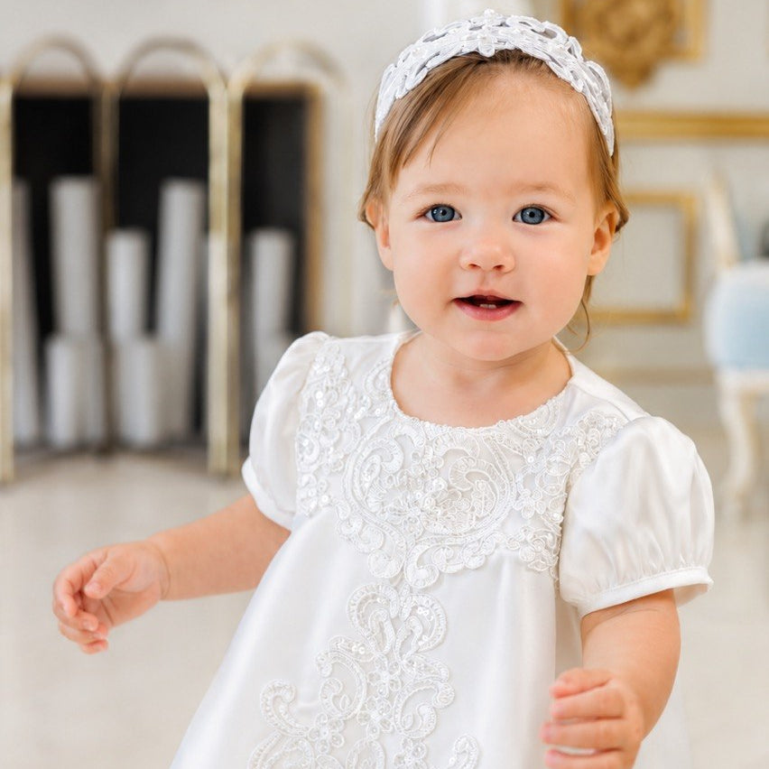 A toddler girl wearing a white baptism dress with short puff sleeves and lace embroidery, along with a matching white lace headband, standing in a bright, elegant room with a blurred fireplace background.