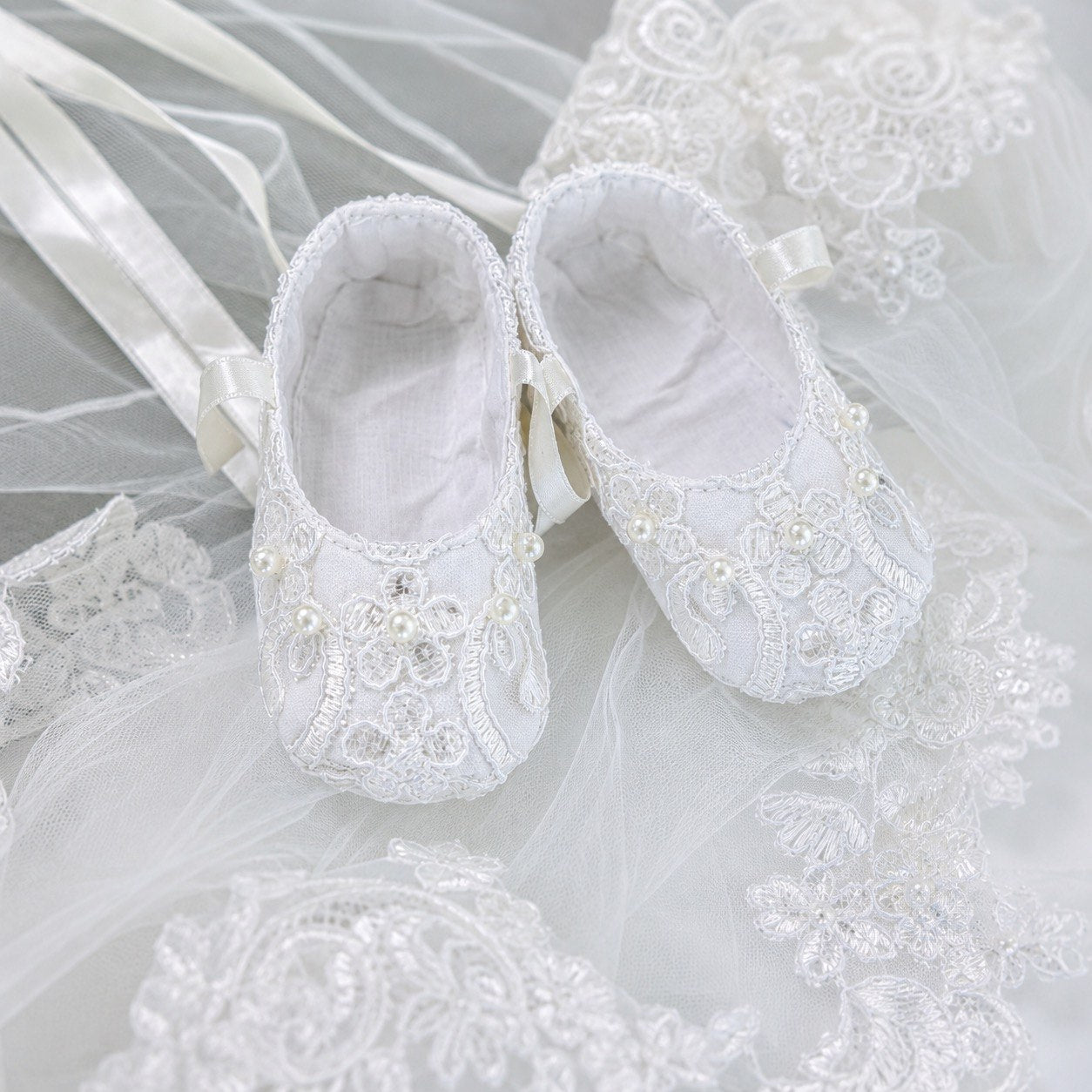A pair of white baby booties with delicate lace embroidery, pearl accents, and satin ribbon ties, styled on a white lace fabric background.