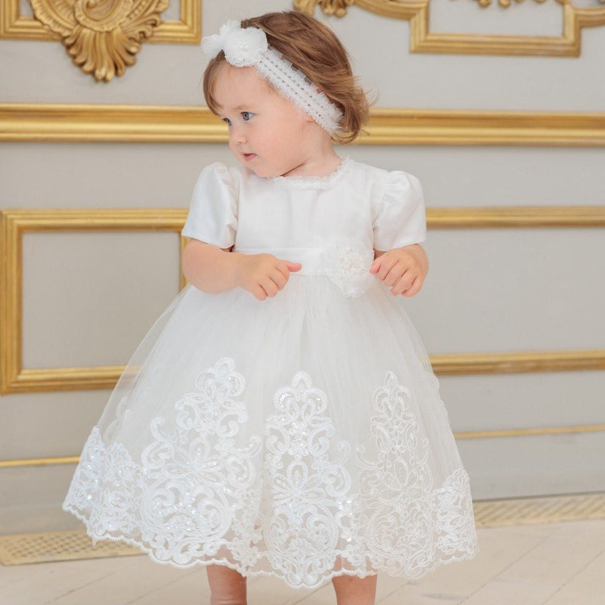 A toddler girl wearing a white short-sleeve lace baptism dress with a matching white headband, standing barefoot in front of an ornate gold and beige wall.