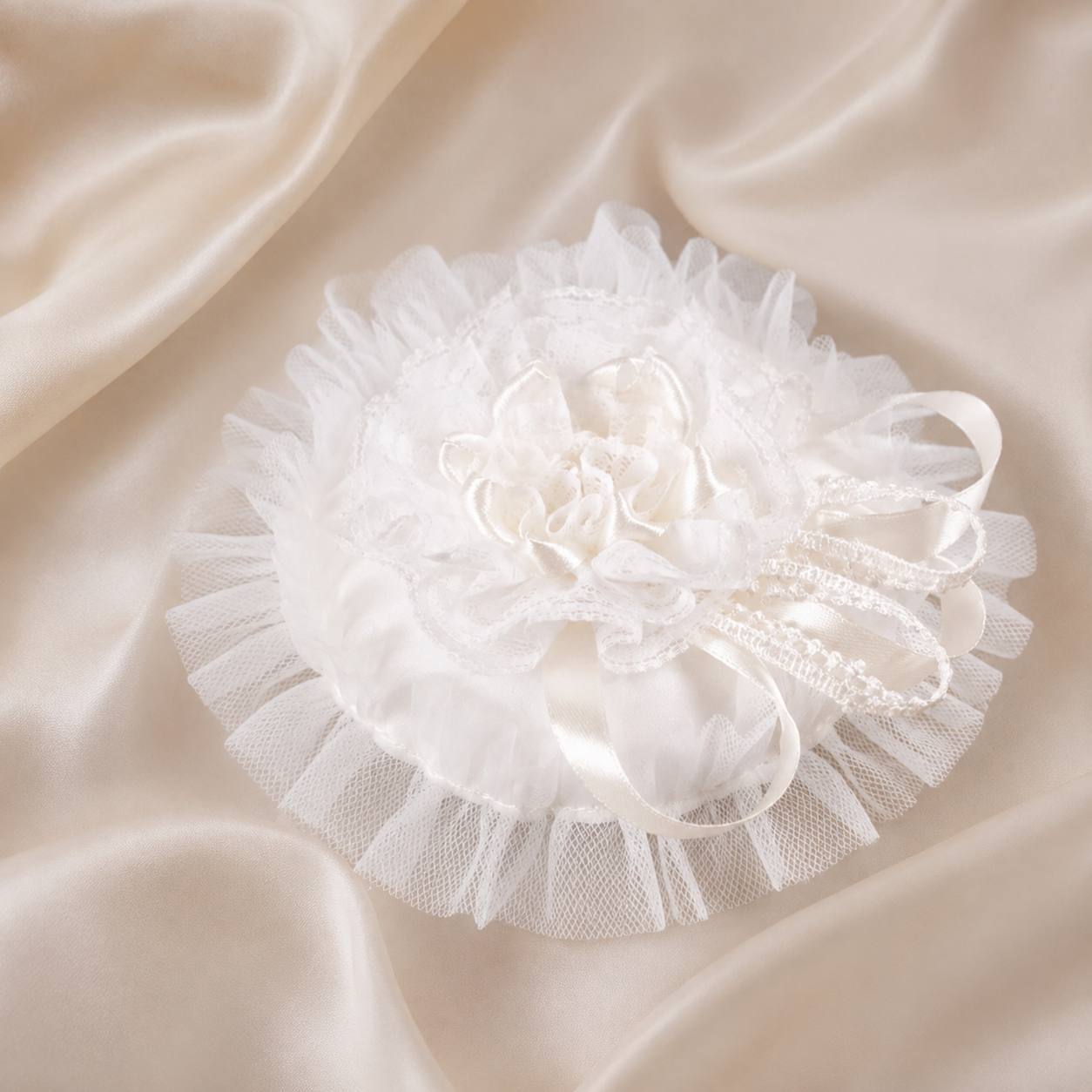 White ruffled lace and satin flower accessory with ribbon details, displayed on a draped cream satin fabric background.