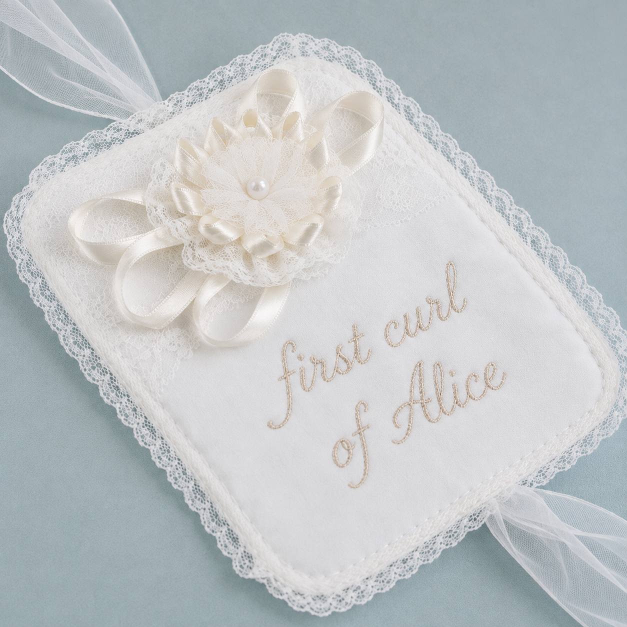 A square white embroidered fabric keepsake tag with lace trim and a white satin flower embellishment. The tag is embroidered with the words 'first curl of Alice' in light beige script and has sheer white ribbon ties at the corners, shown on a soft dusty blue background.