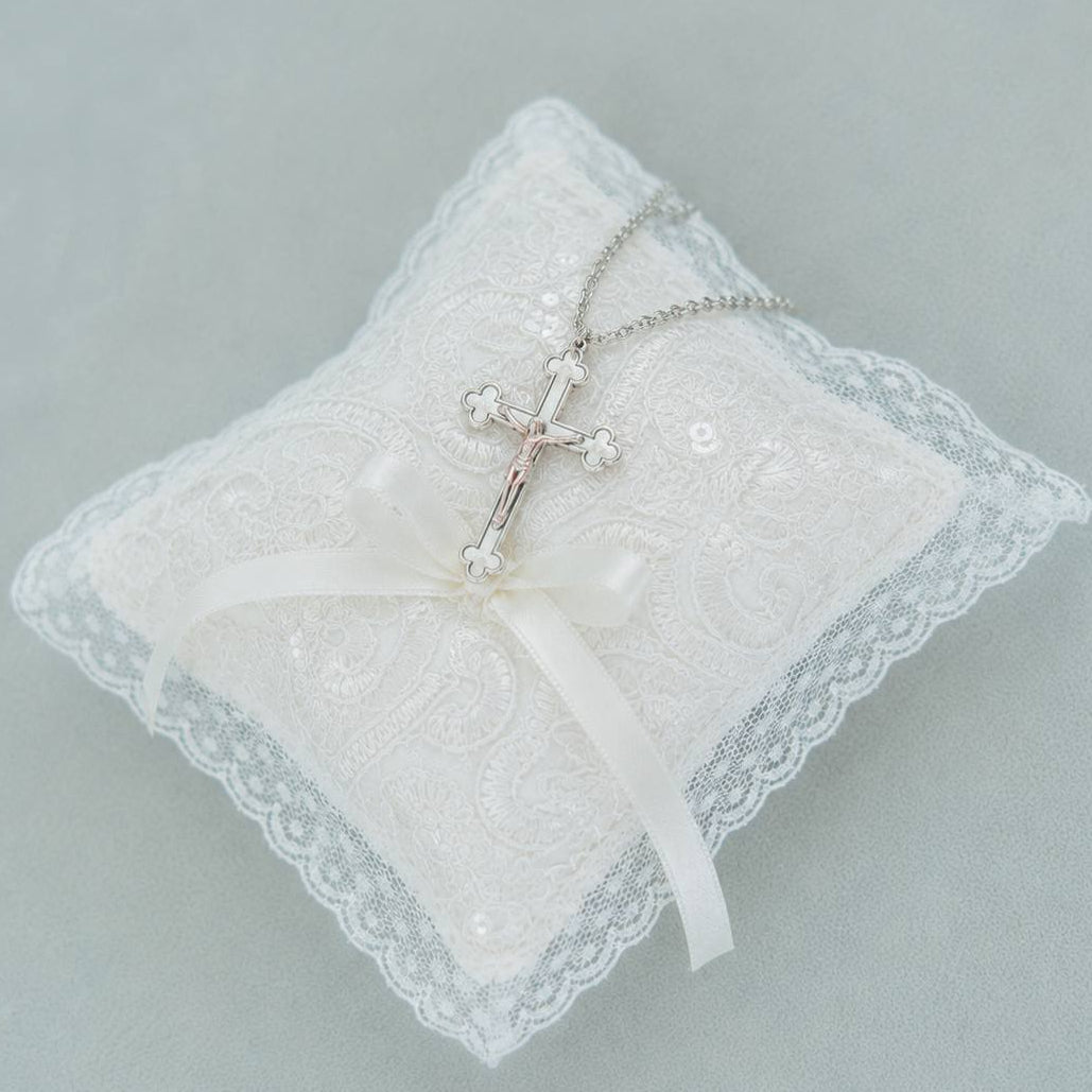 A silver crucifix pendant necklace displayed on a white lace ring pillow with a white satin ribbon, set against a soft gray background.