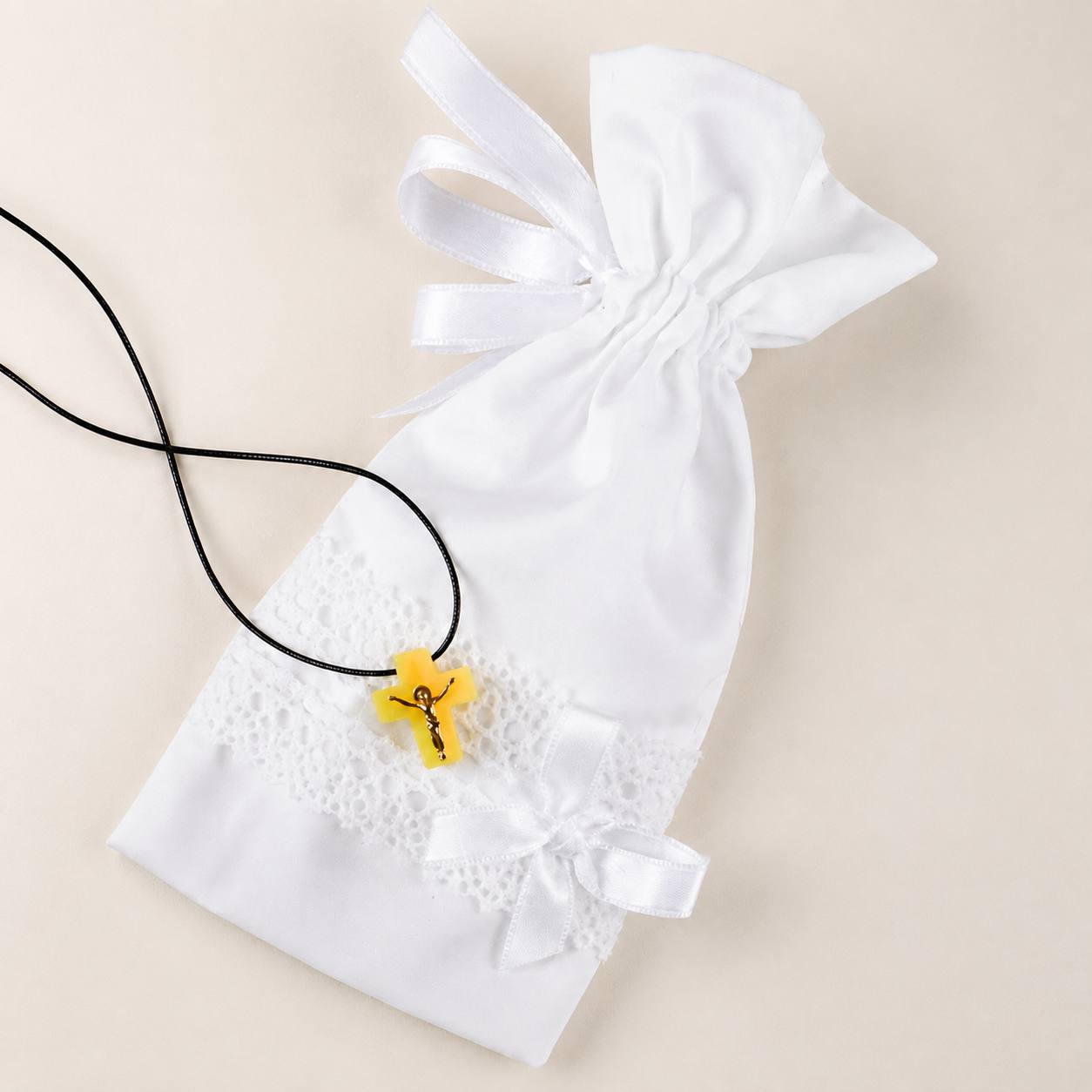 A white satin drawstring pouch with lace trim and ribbon bows, laid on a light beige background next to a black cord necklace with a small yellow cross pendant.