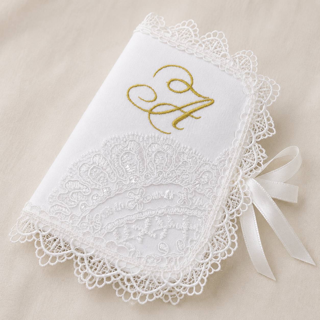 A white embroidered monogrammed keepsake album or notebook with delicate white lace trim, a scalloped edge, and a white satin ribbon bow, displayed on a neutral beige fabric background.