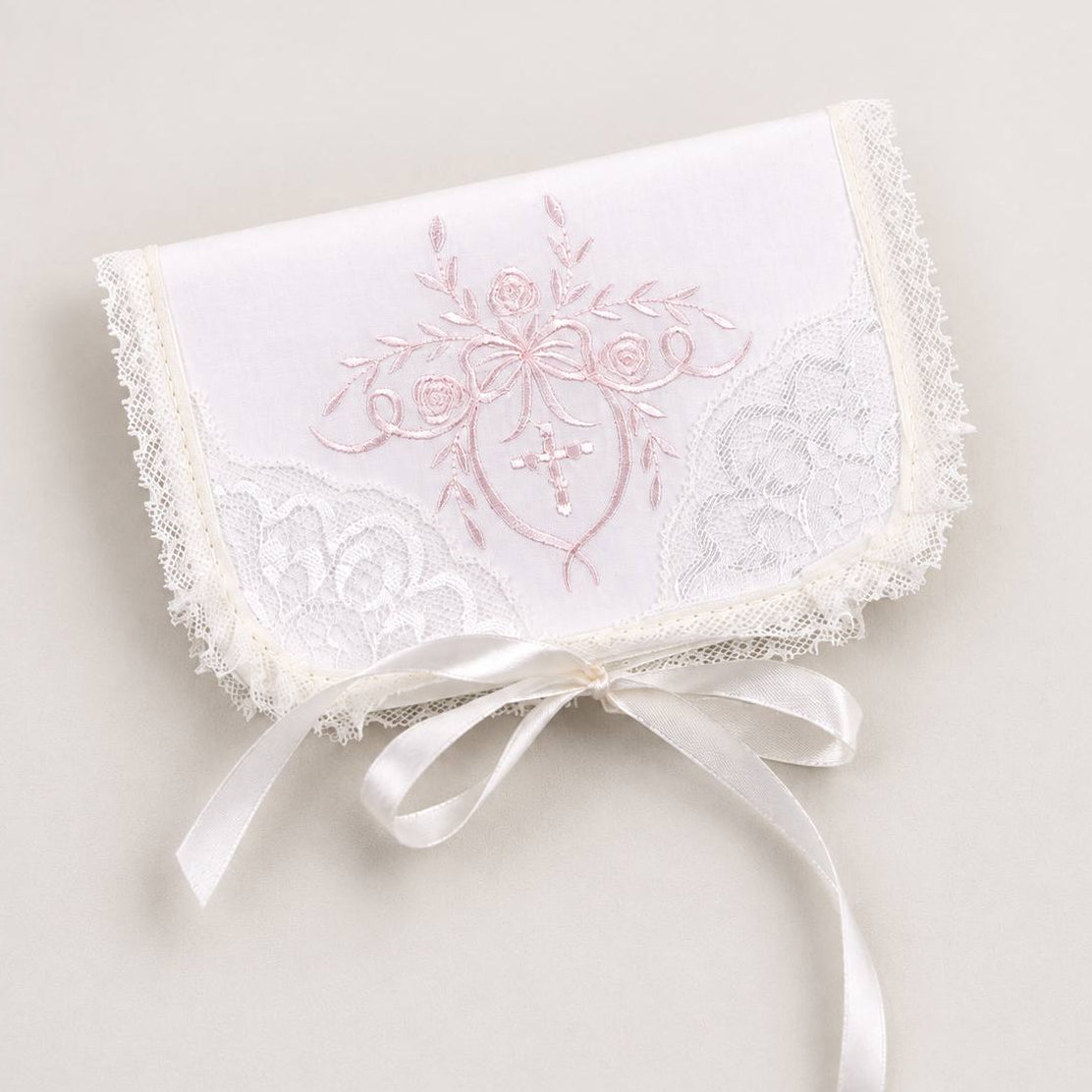 A white embroidered lace pouch with a satin ribbon bow, featuring a pink floral design and a small cross on the front, set against a light beige background.